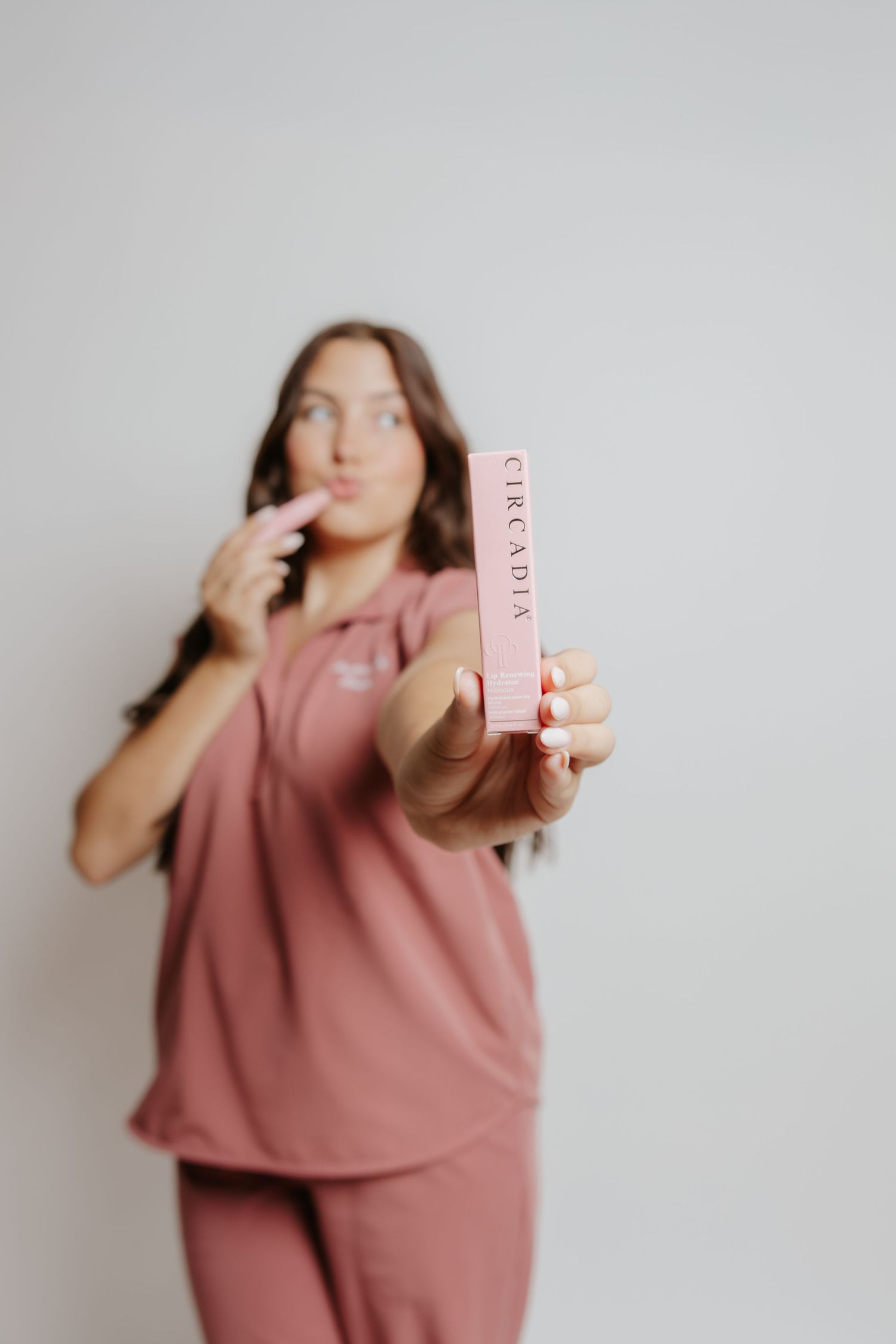 Woman in pink scrubs holding Circadia skincare product while applying lip treatment against white background.
