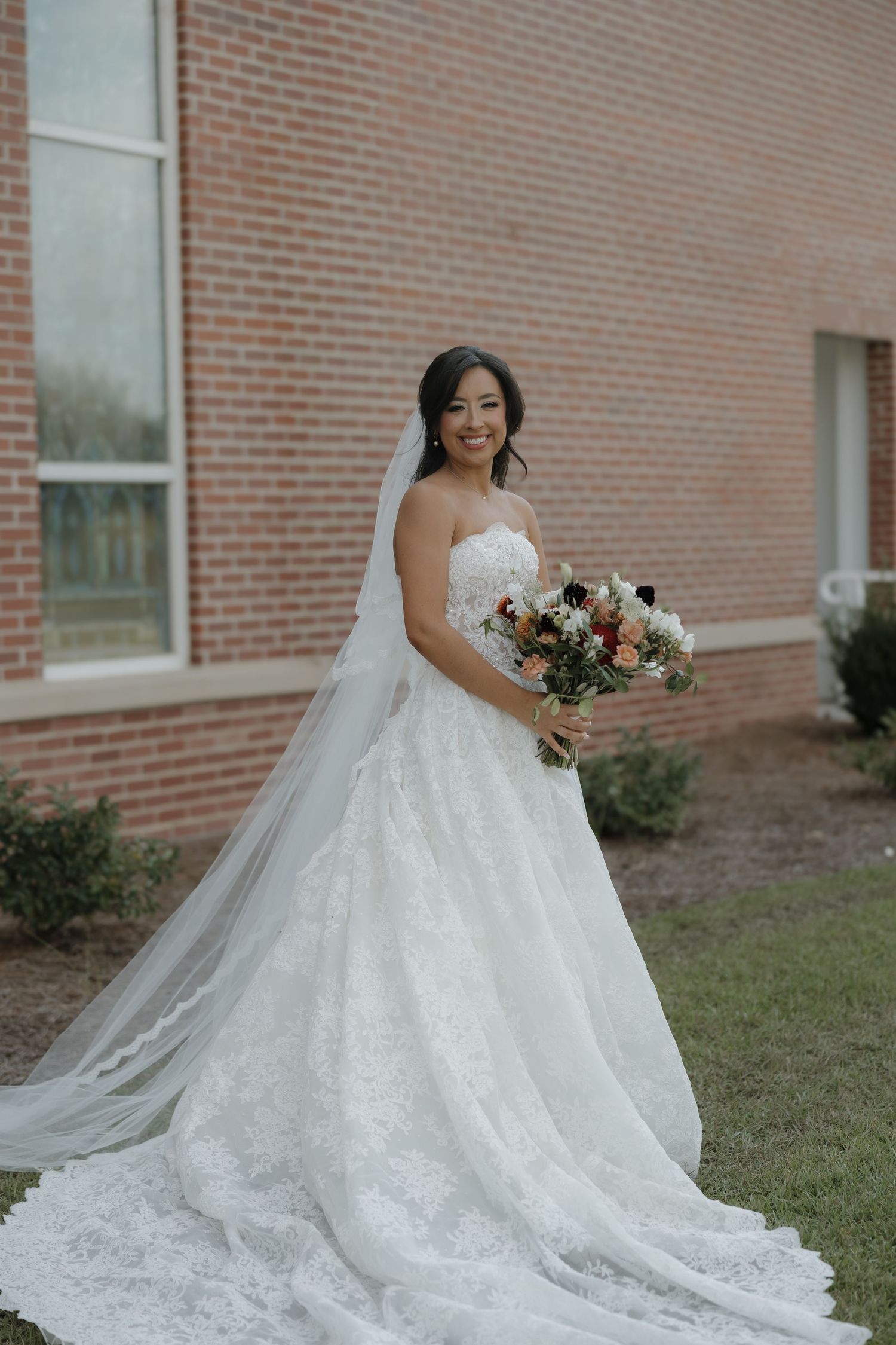 A smiling bride in a lace ball gown and long veil holds a colorful bouquet while standing on grass beside a brick building.