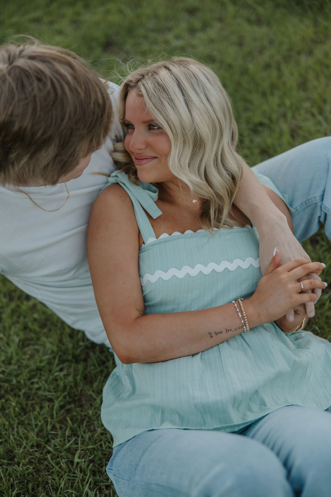 Couple sitting on green grass, woman in mint smocked top smiling as man whispers in her ear.