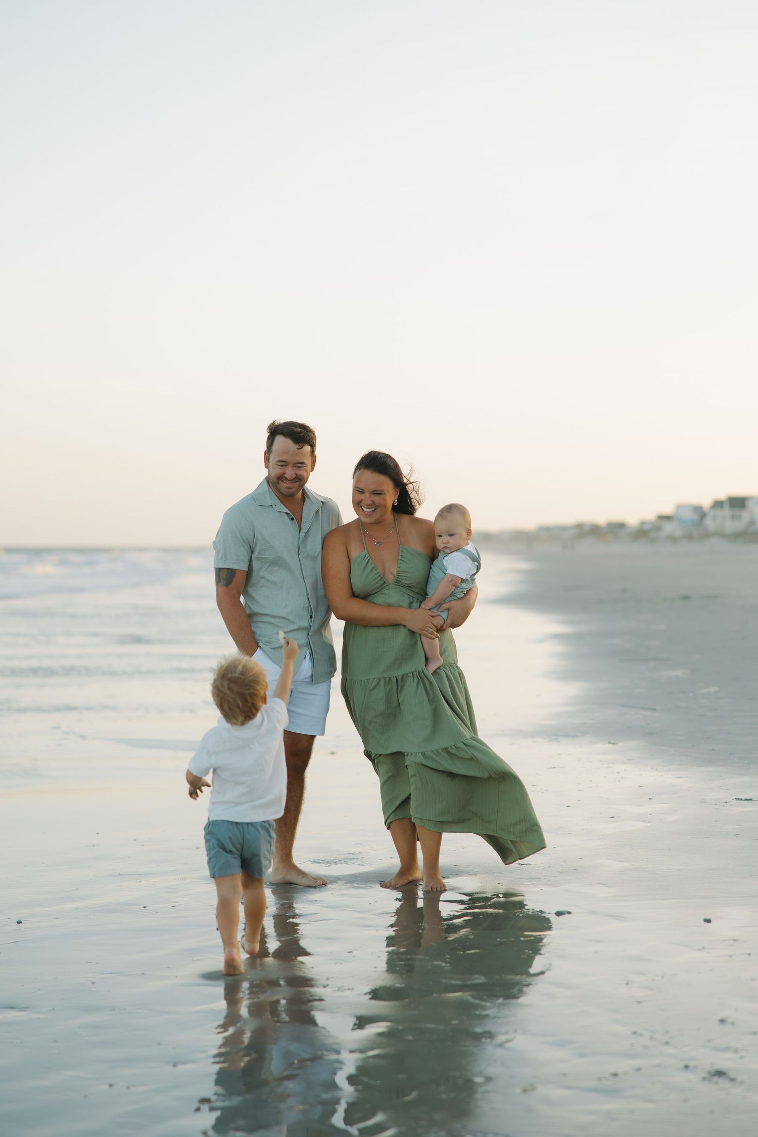 Family of four enjoying a sunset beach walk, mother in green dress holding baby while toddler runs toward the water.