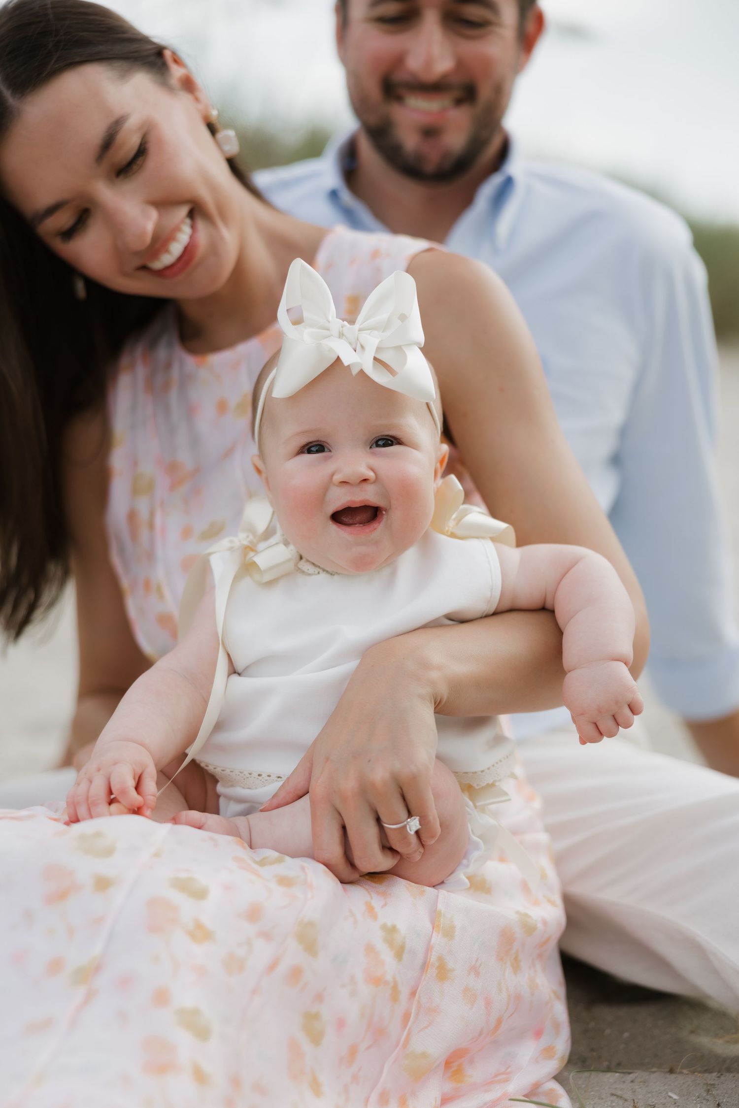 Happy family with smiling baby girl in white bow headband sitting outdoors during beach photo session.