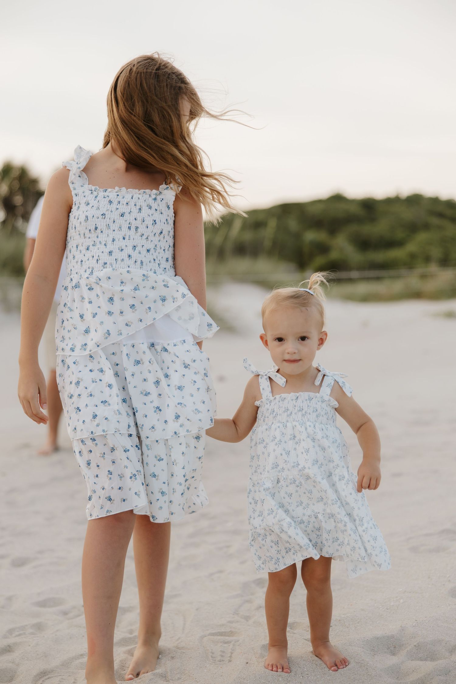 Two young sisters in matching floral dresses holding hands on a sandy beach at sunset.