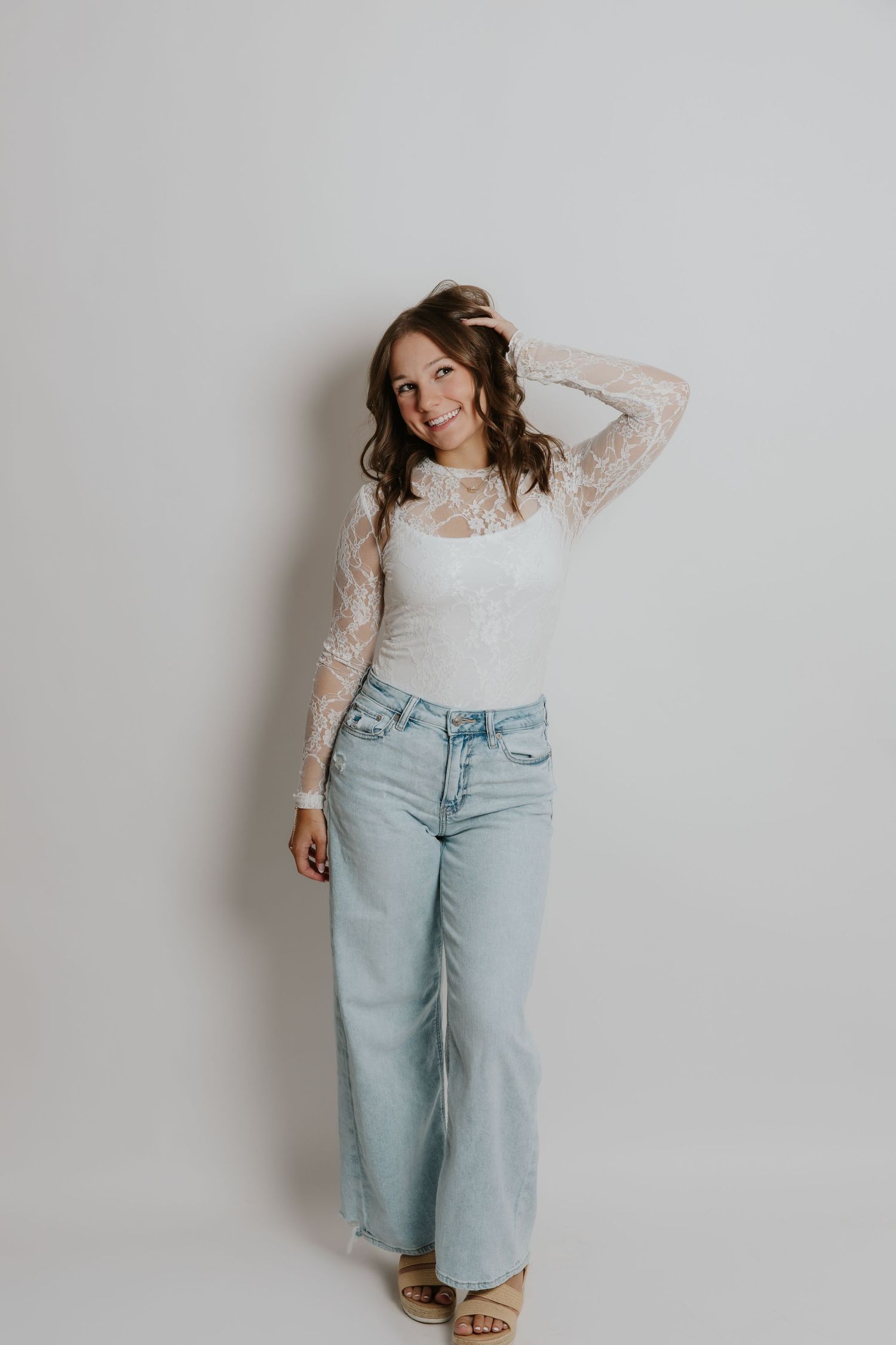 Happy woman in white lace bodysuit and wide-leg light wash jeans leaning against white studio wall.