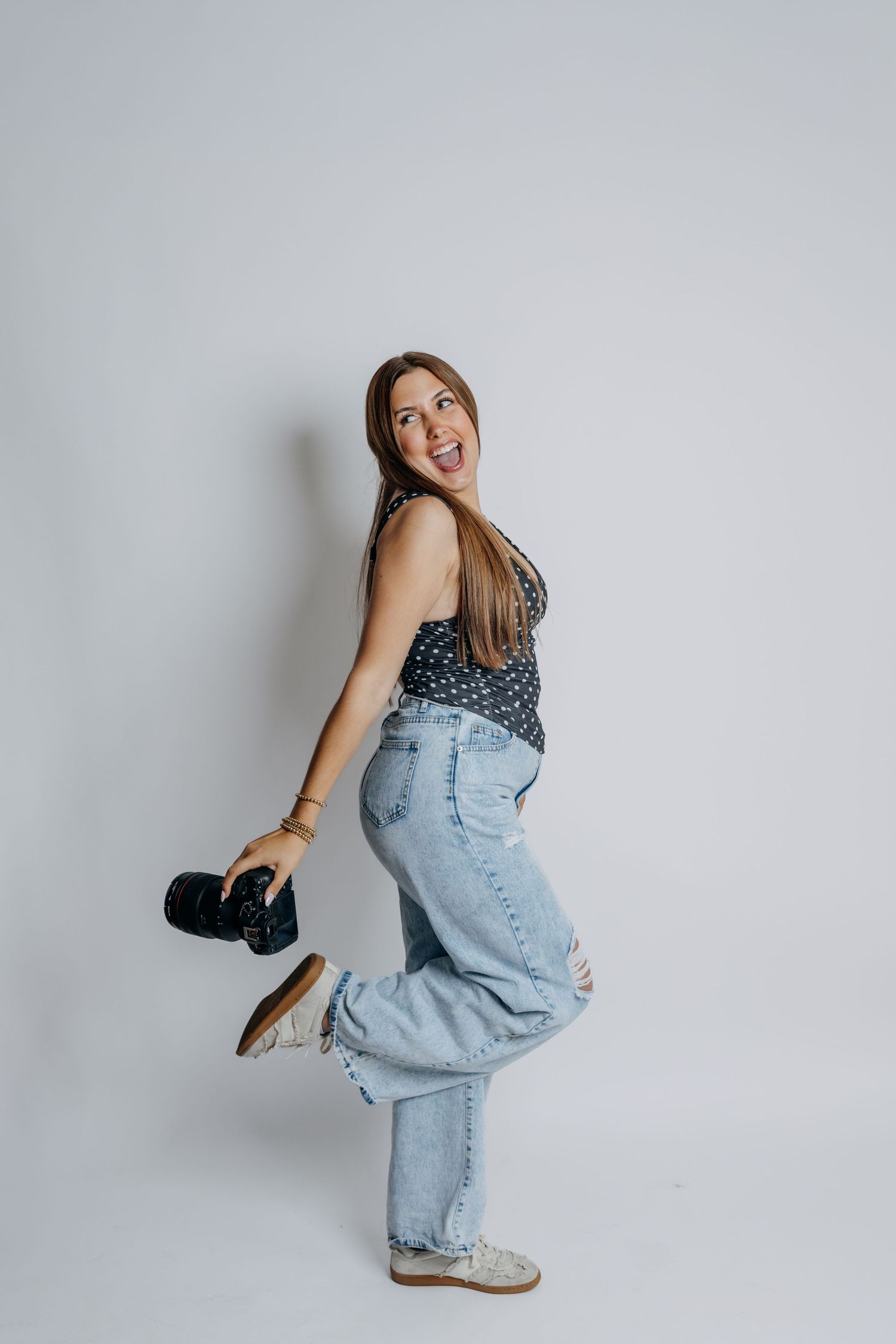 A cheerful woman in polka dot top and jeans holds a camera while playfully posing against a white background.