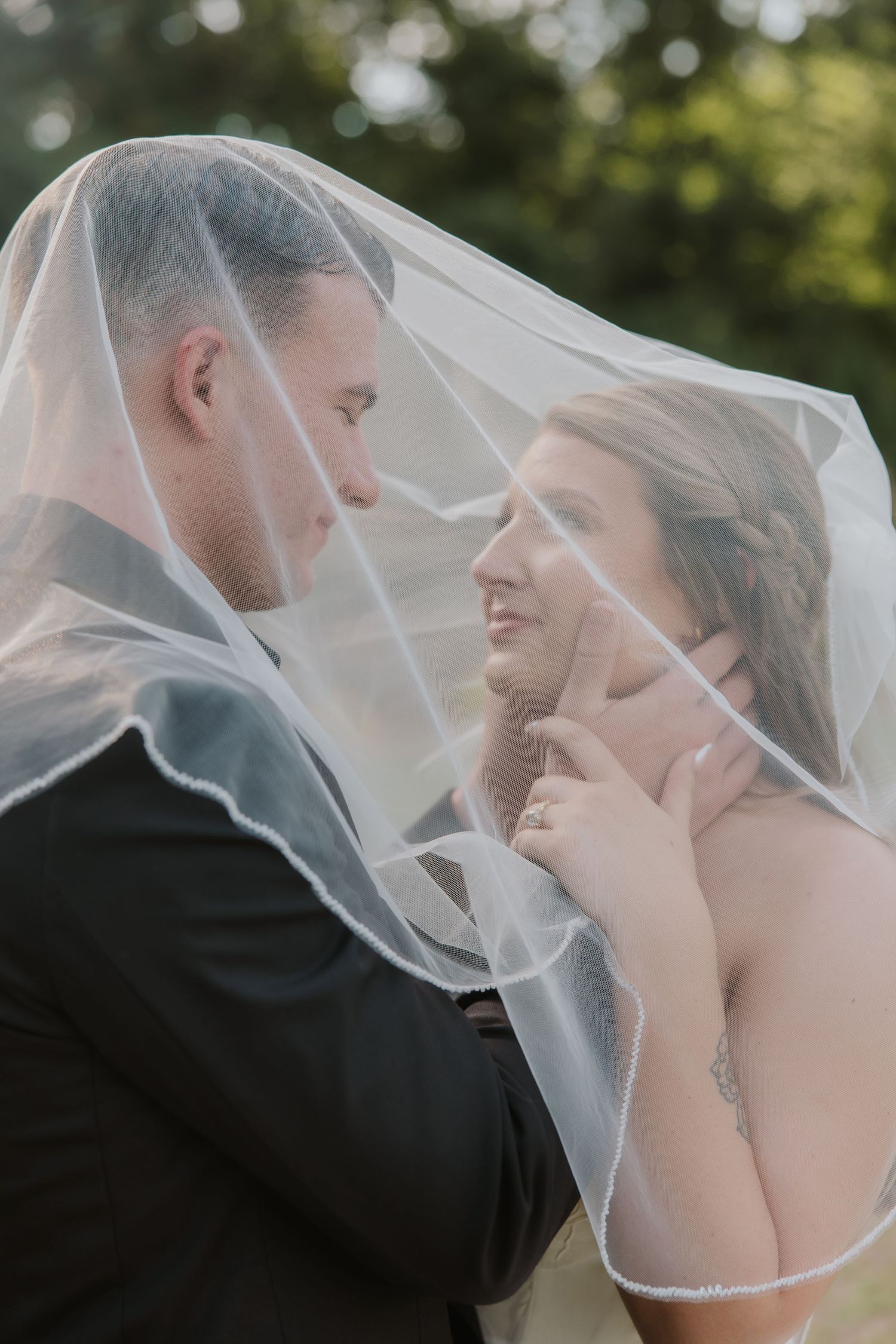 Bride and groom share an intimate moment under a flowing white veil outdoors on their wedding day.
