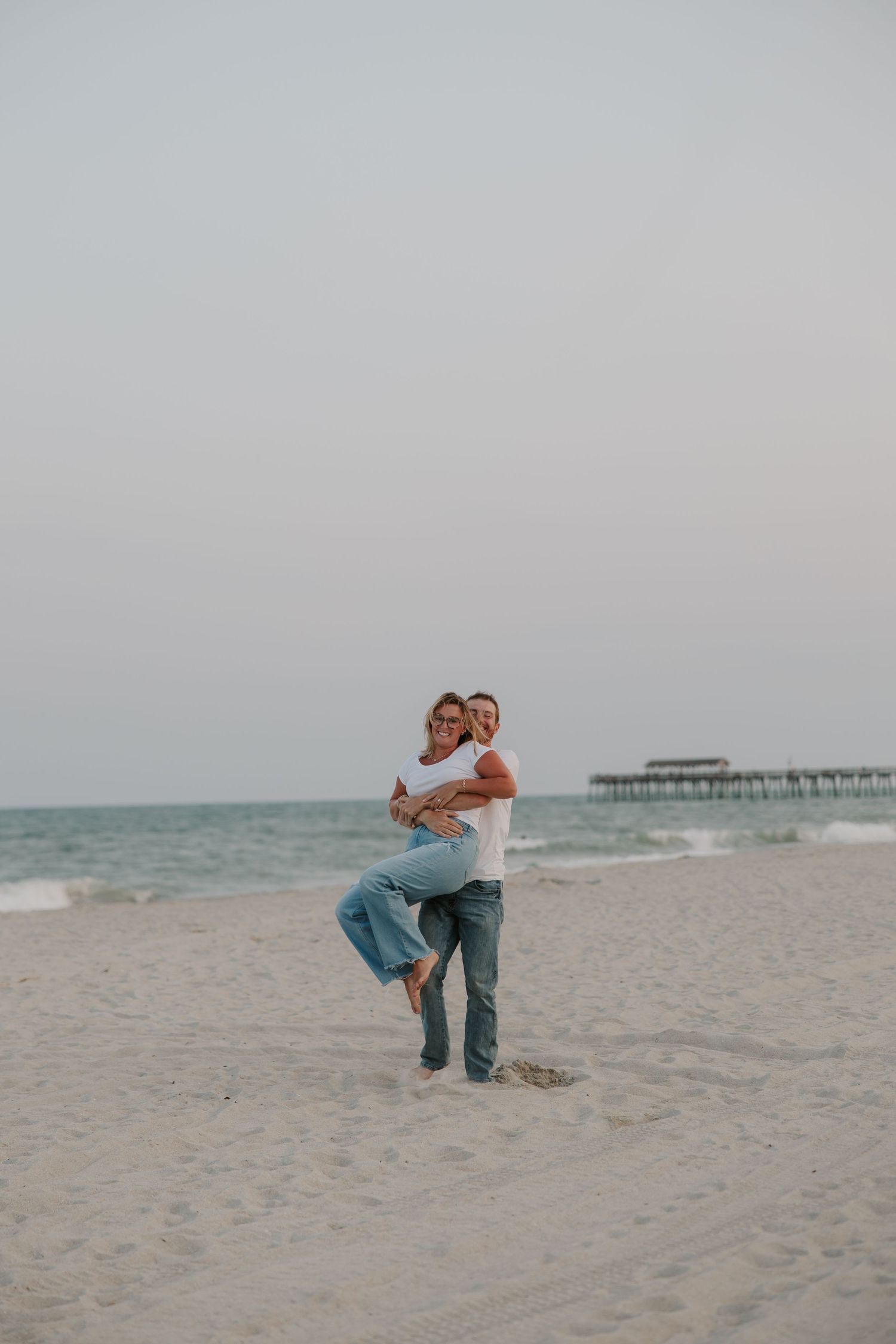 Couple embracing on a sandy beach near a pier, man lifting woman in white top and jeans on overcast day.