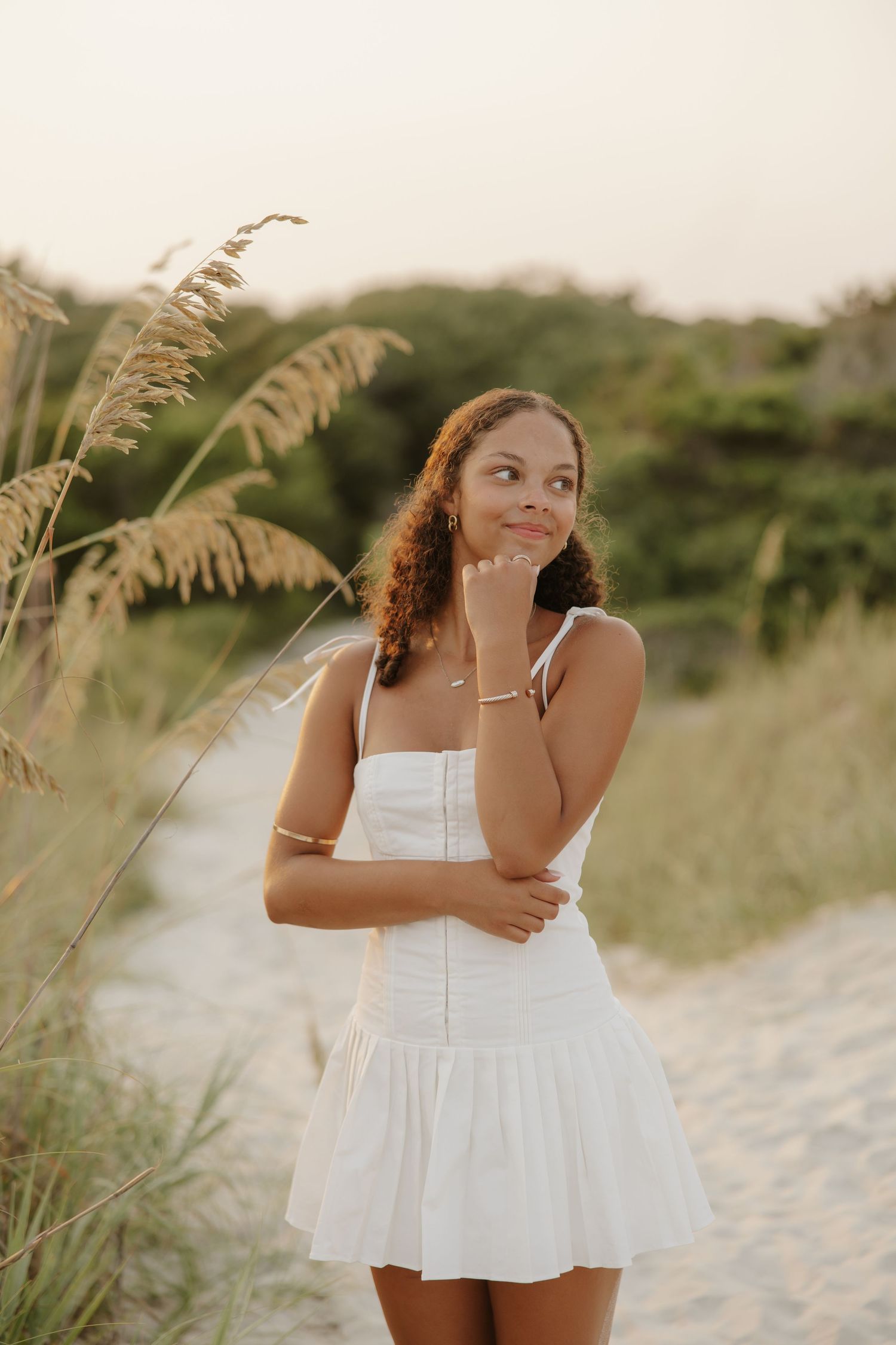 Young woman in white sundress standing on sandy beach path surrounded by sea oats at sunset.