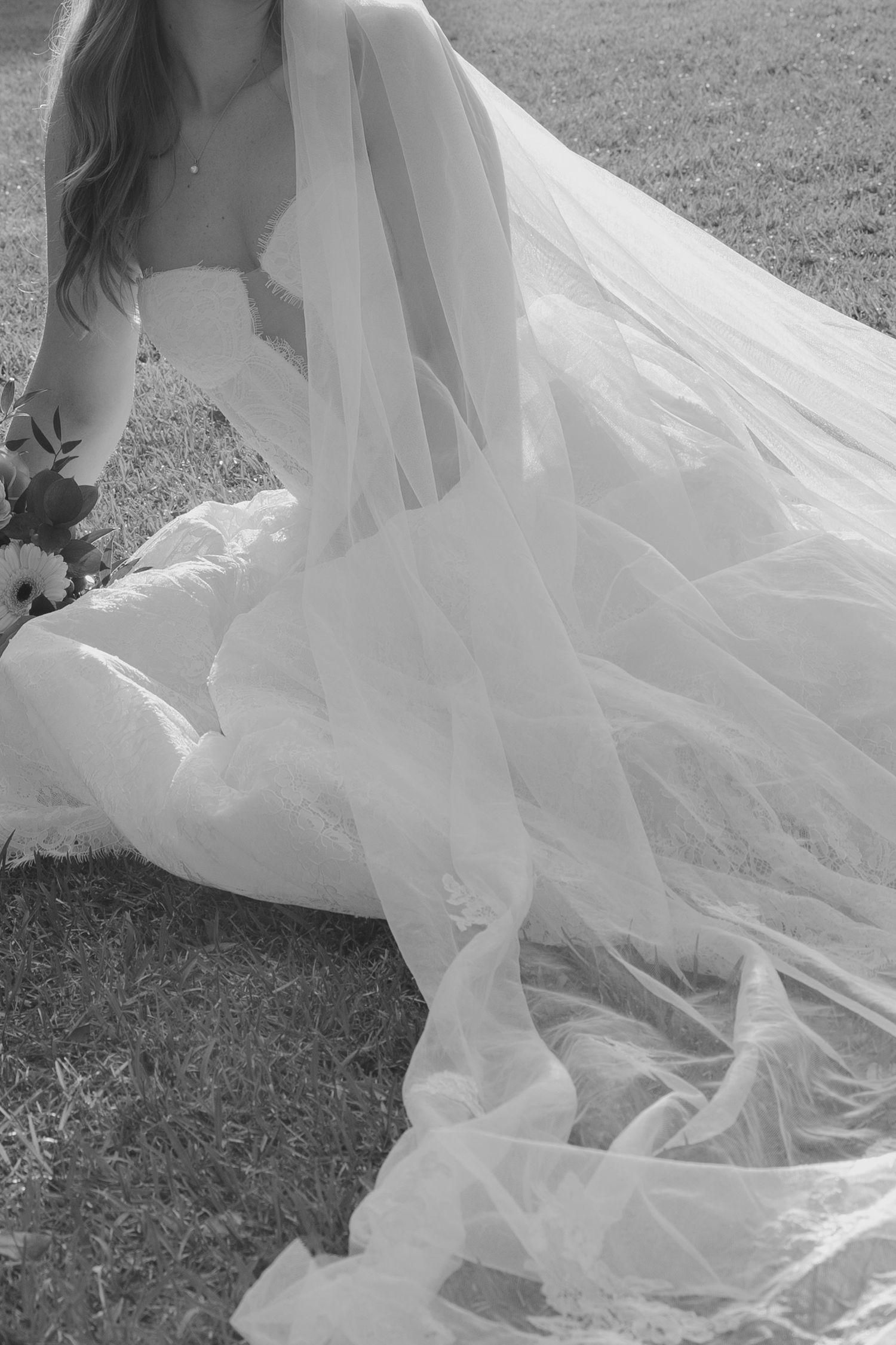 Black and white photo of bride sitting in grass with flowing tulle wedding dress.