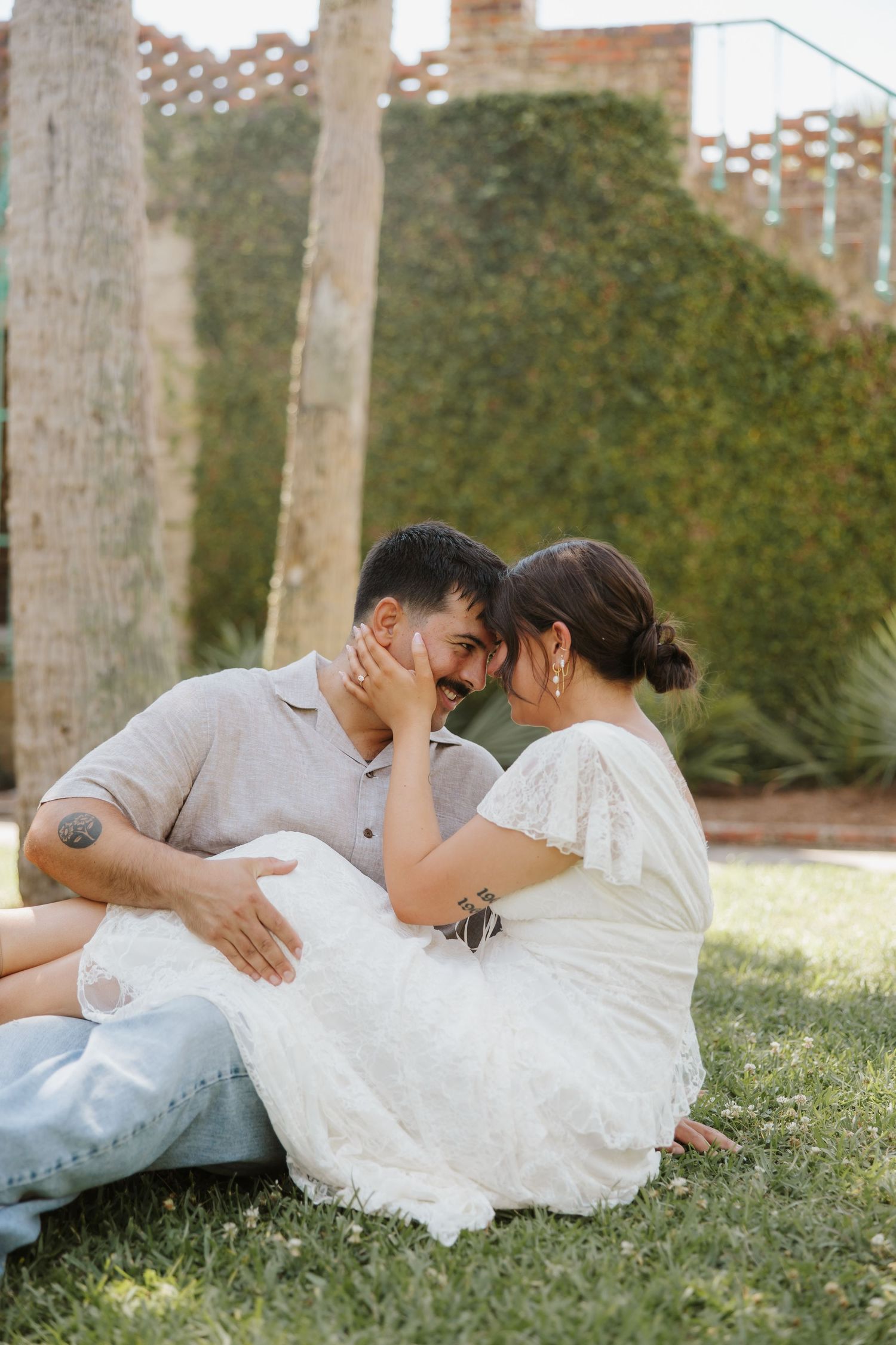Couple sitting on grass in a garden, sharing an intimate moment, woman in white dress touching man's face.