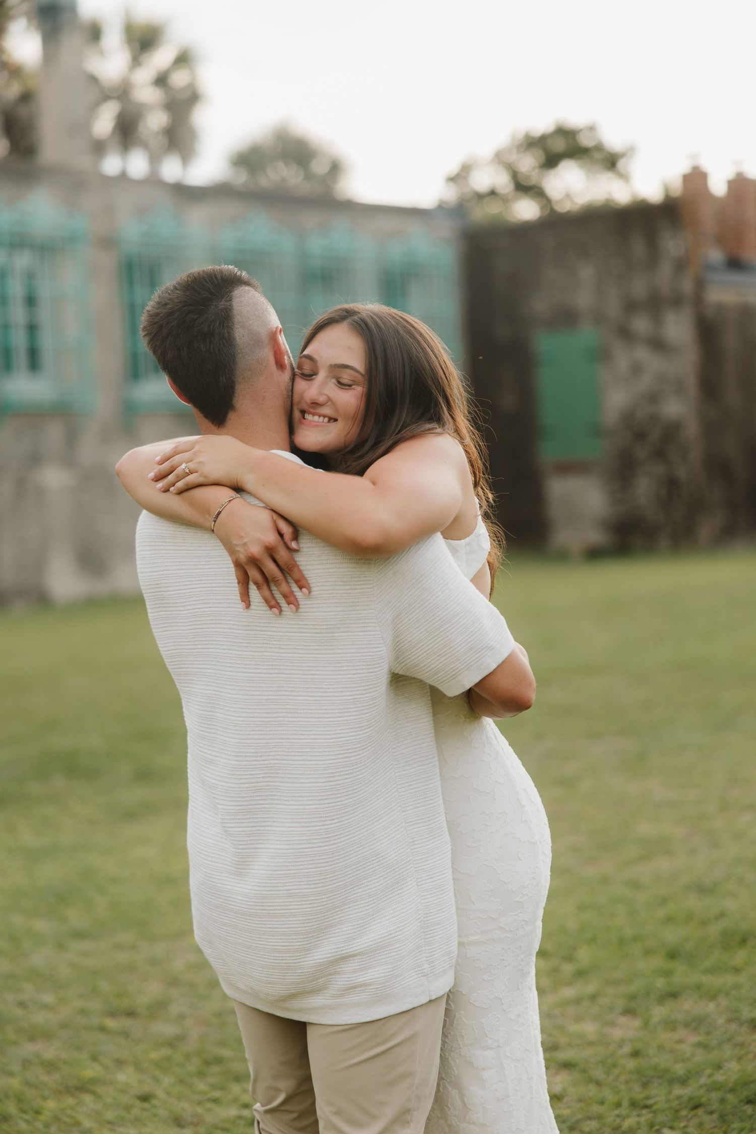 Couple embracing and smiling outdoors near historic stone building with teal windows on green lawn.