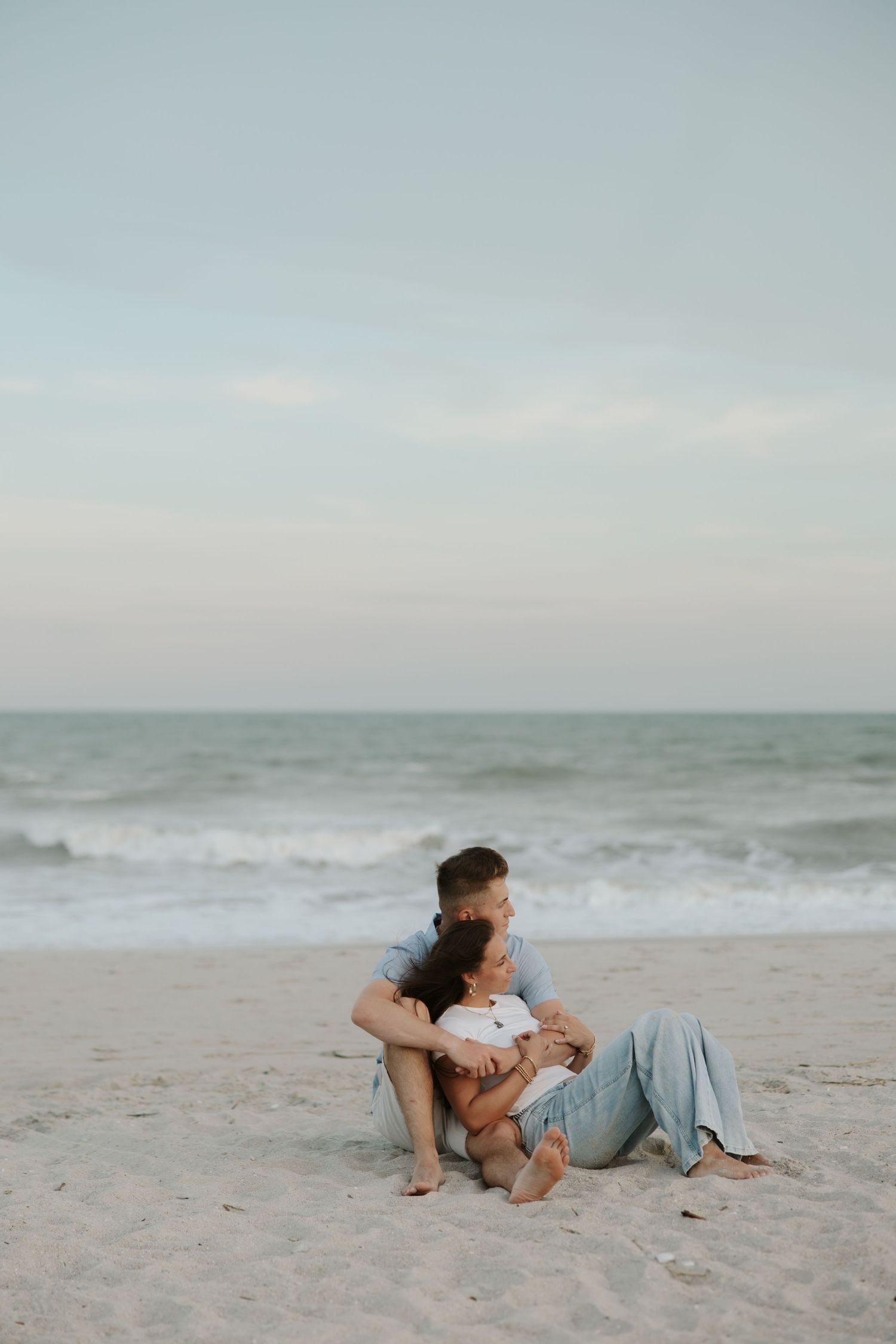 Couple sitting together on a sandy beach at dusk, embracing with ocean waves in the background.