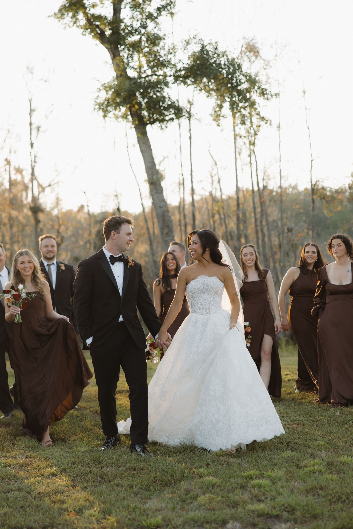 Bride in a strapless lace gown holds hands with the groom in a black tuxedo as bridesmaids in brown dresses walk behind them outdoors.