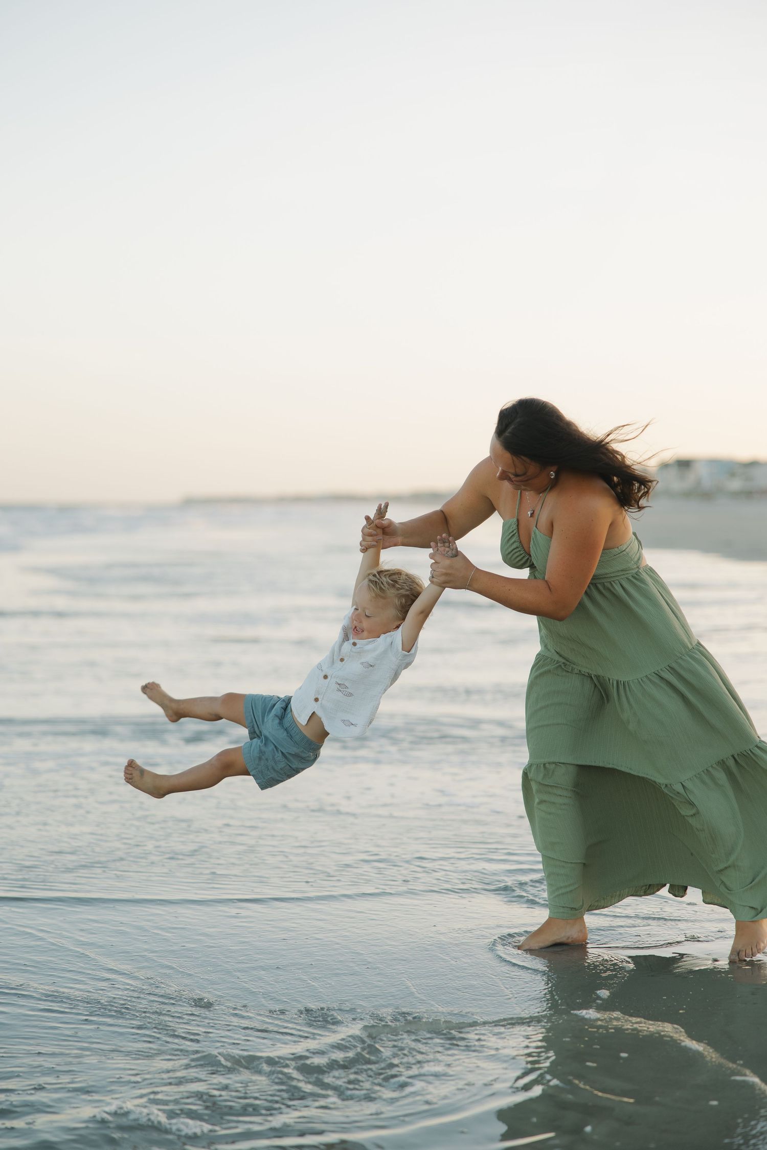 Mother in green dress swinging toddler boy over ocean waves on sandy beach at sunset.