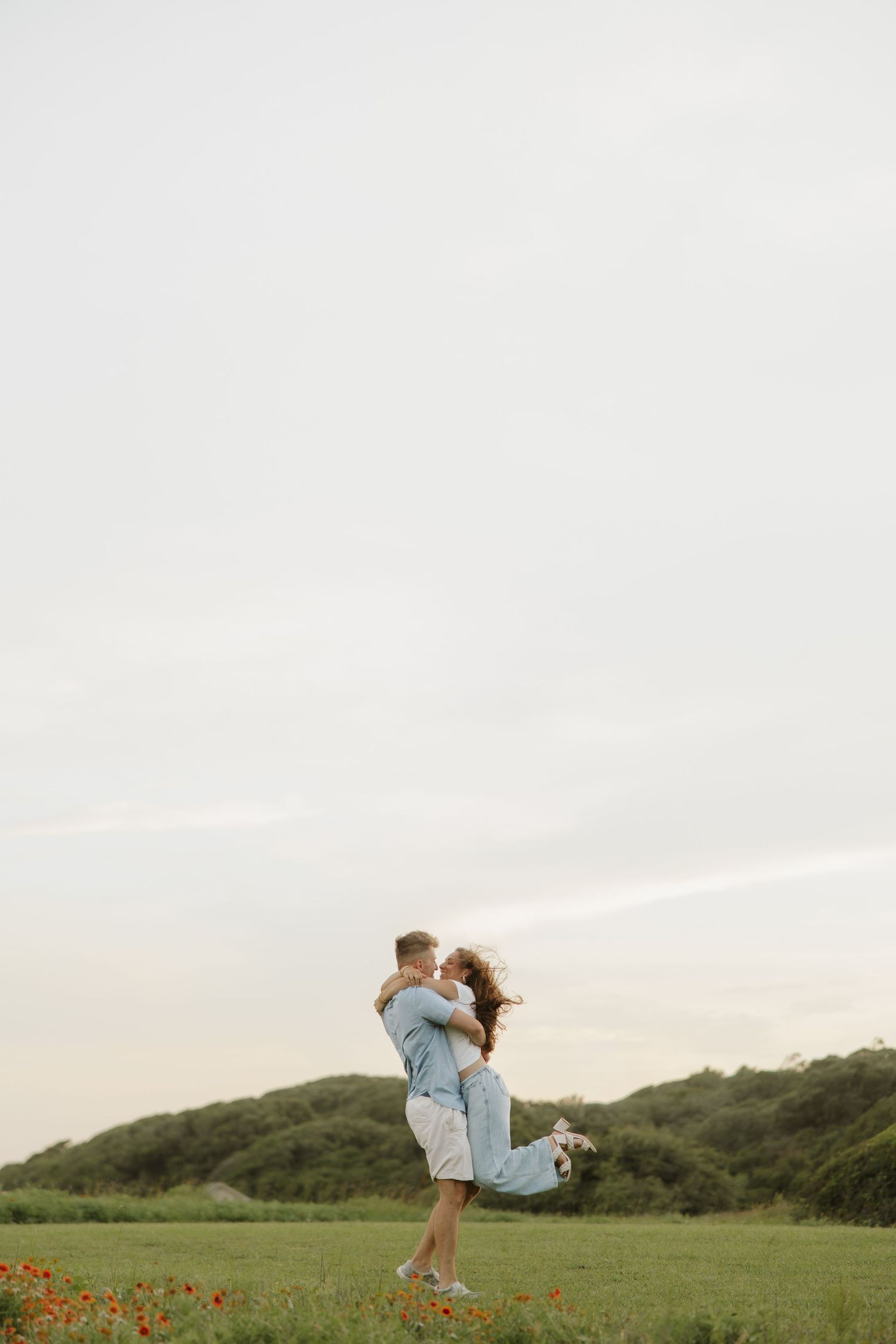 Couple embracing in a green field, man lifting woman while she holds a bouquet, with hills and cloudy sky behind them.