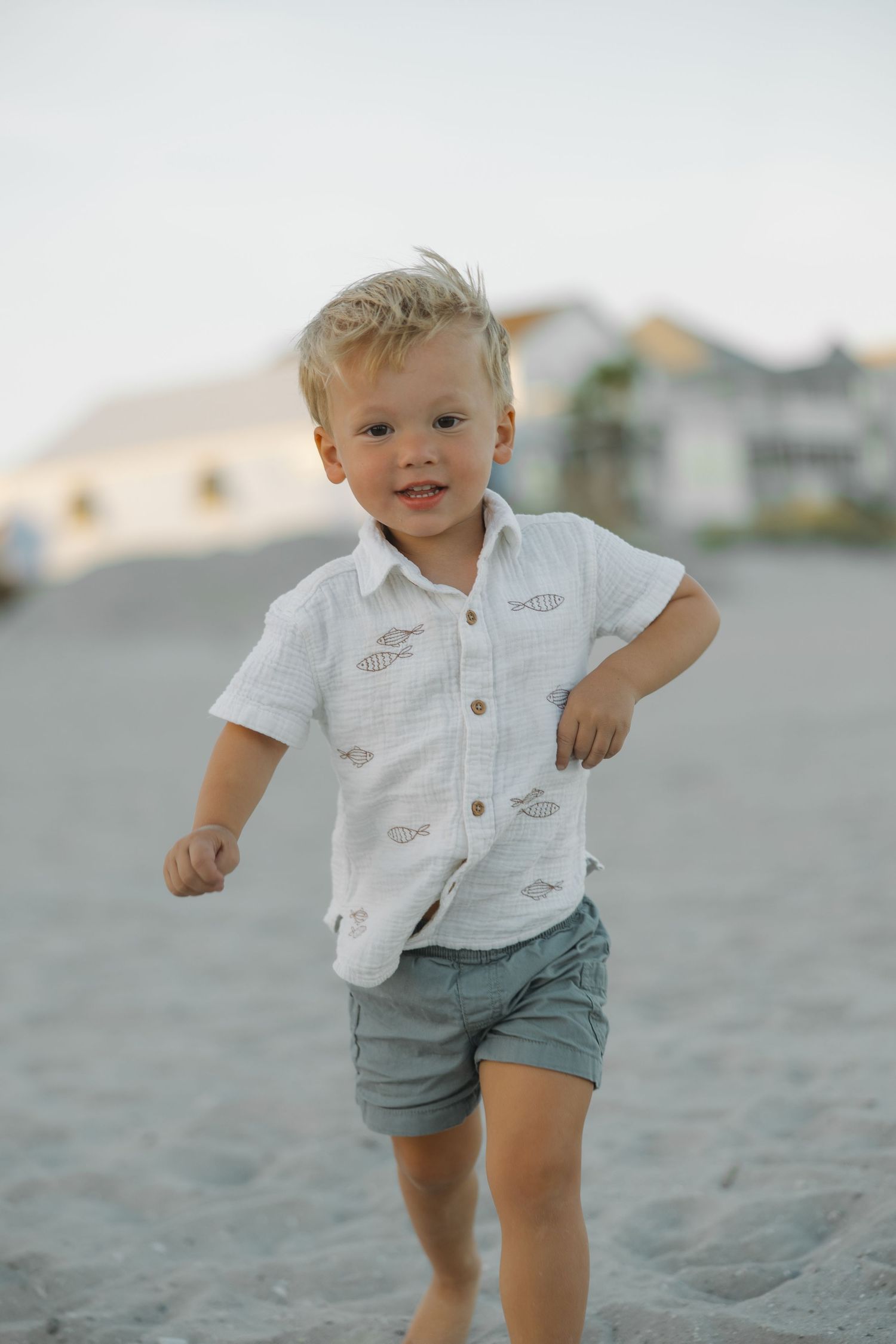 Smiling toddler boy in white fish-print shirt and gray shorts running on a sandy beach.