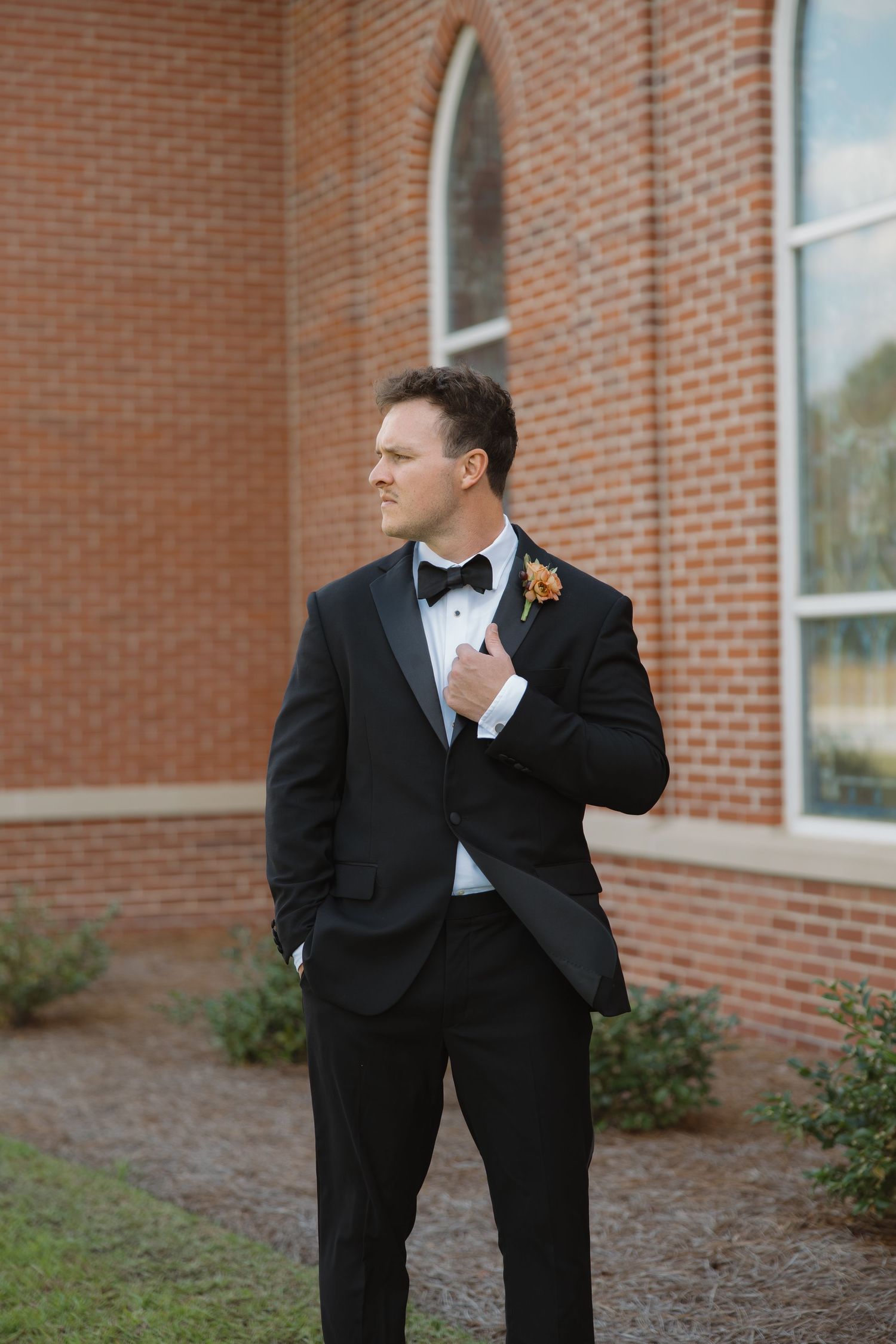 A groom in a black tuxedo and bow tie with an orange boutonniere poses beside a red brick building.