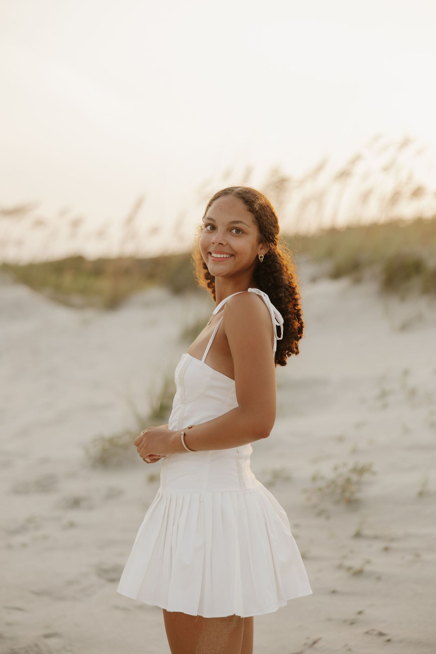 Young woman in white sundress smiling on sandy beach dunes at golden hour sunset.