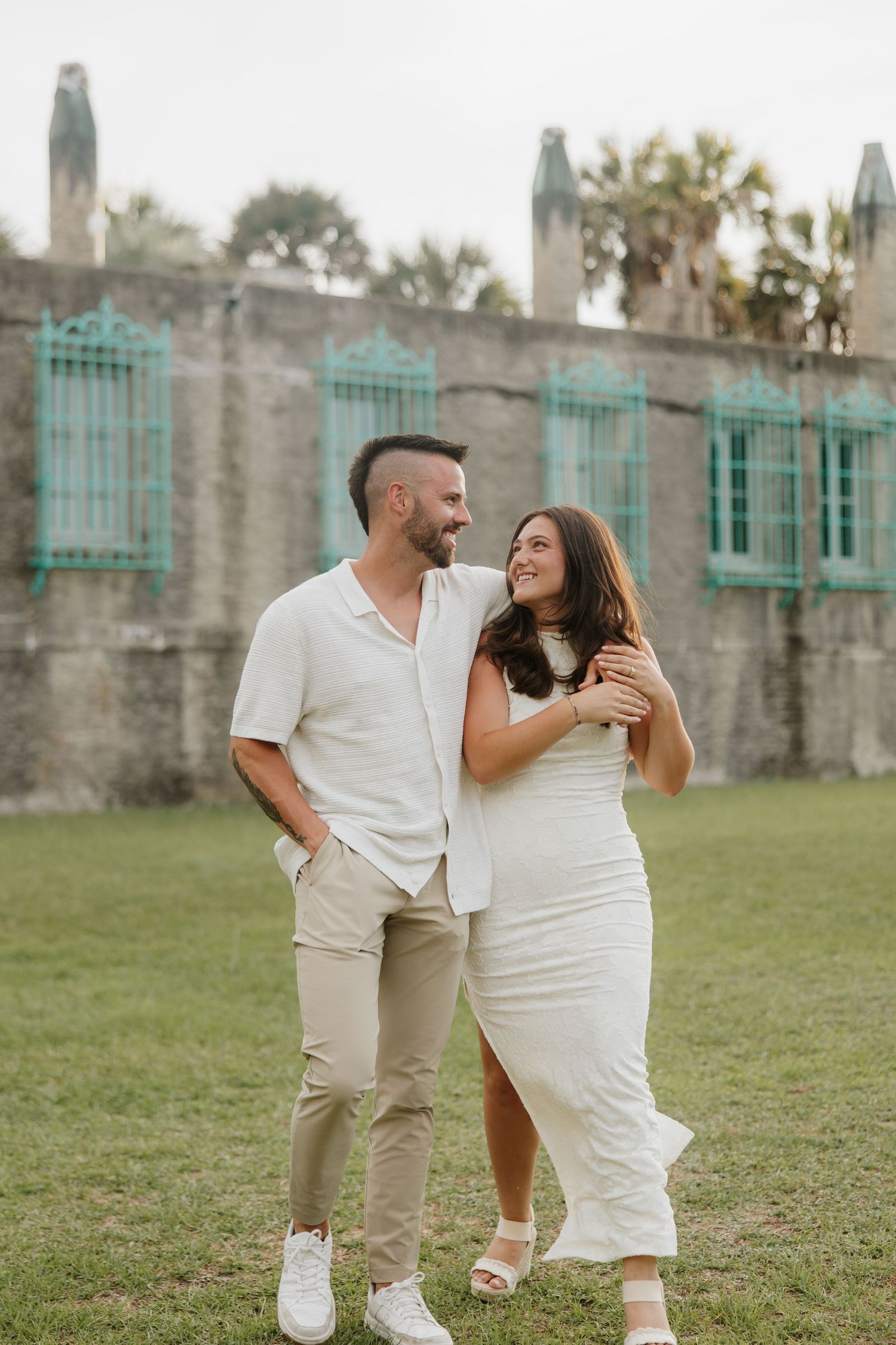 A couple in matching white outfits smiles and embraces on a lawn before a historic stone wall with teal windows.
