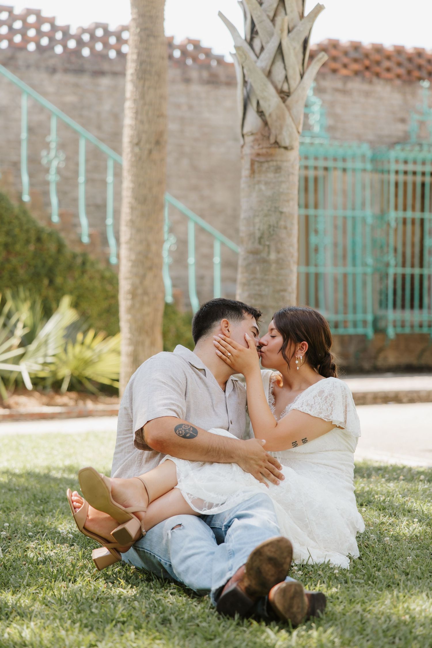 Couple sitting on grass sharing a kiss, woman in white lace dress, man in light shirt and jeans with tattoo.