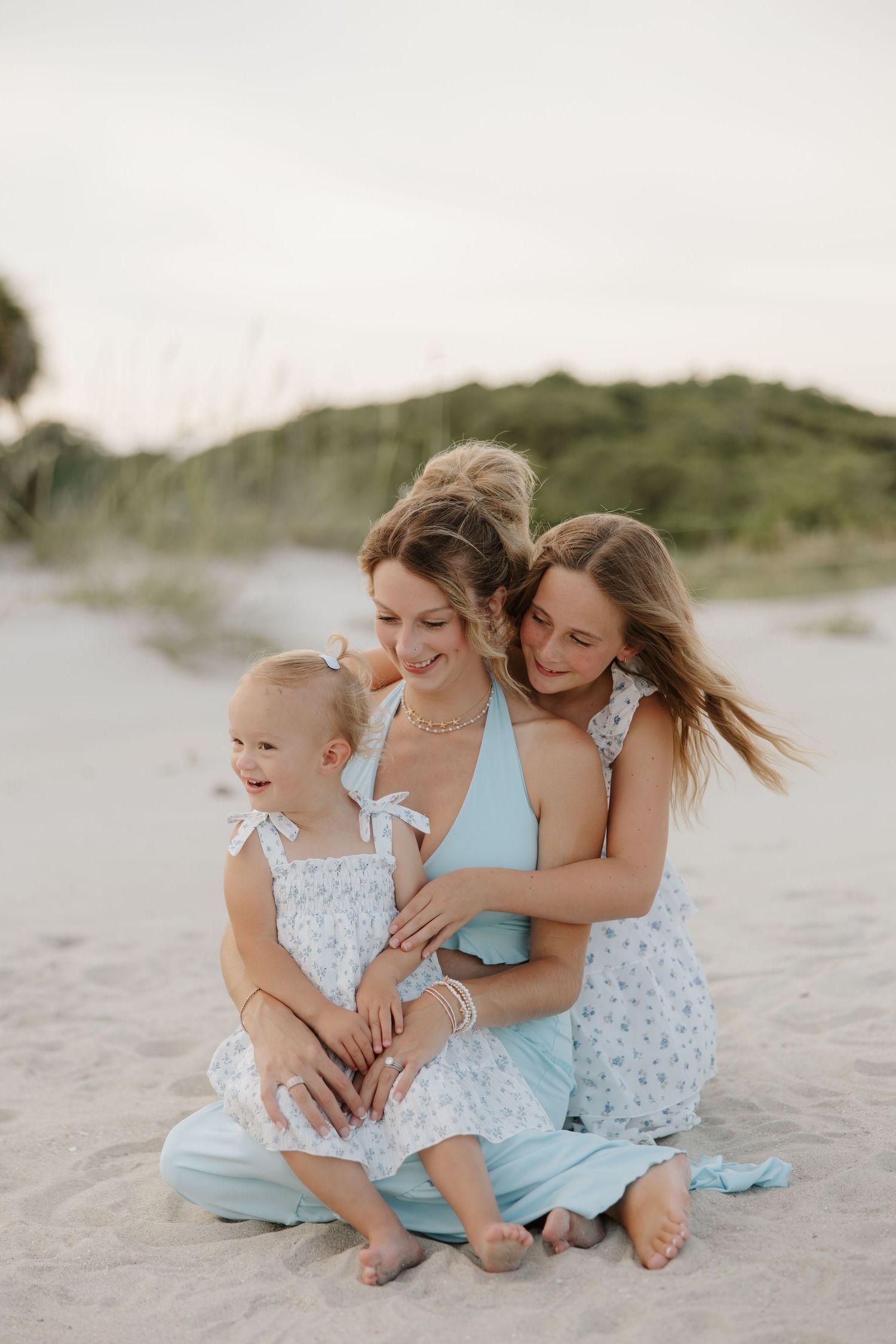 A mother and two daughters in matching blue floral outfits sit together on a sandy beach, smiling and embracing warmly.