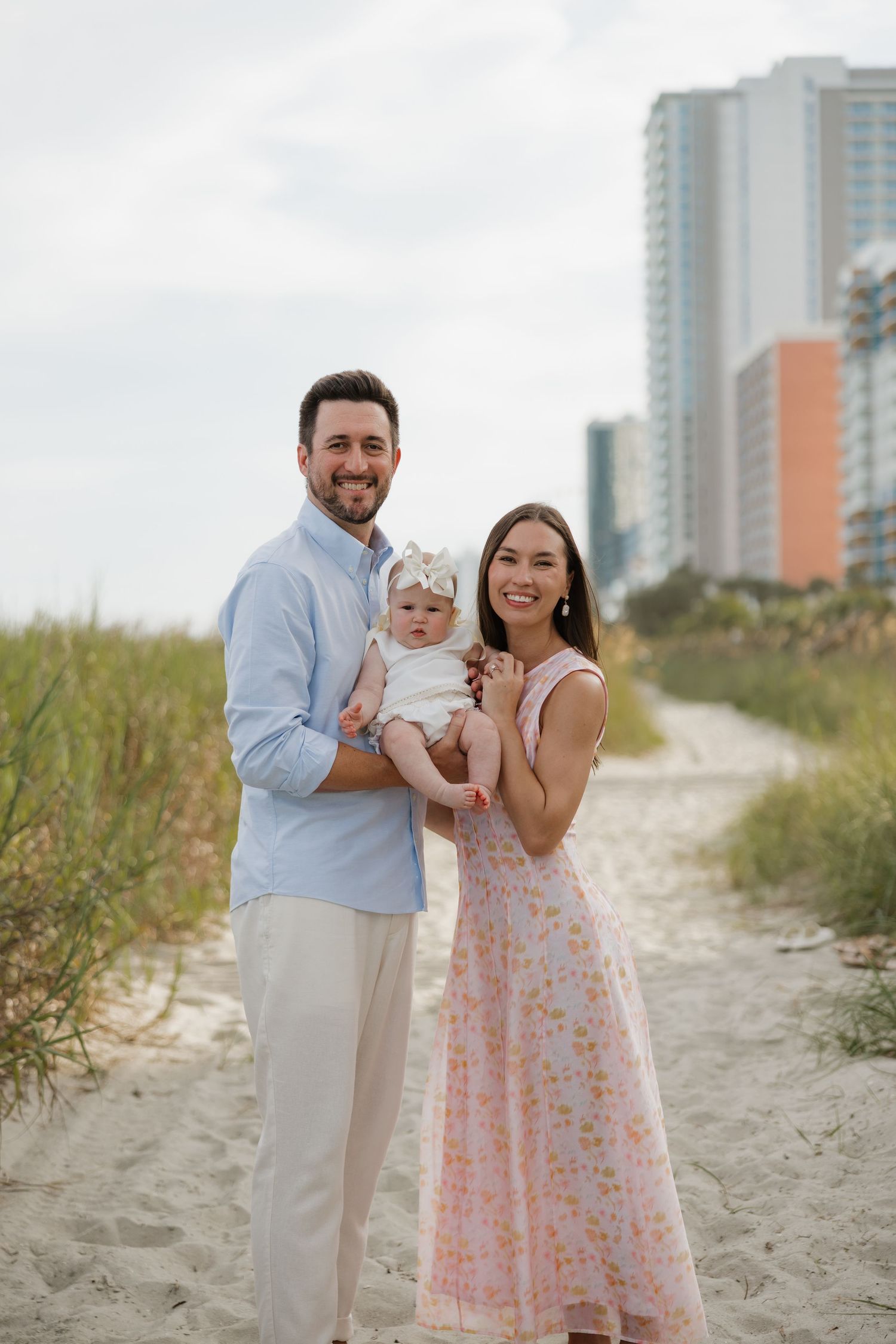 A happy couple holding their baby girl poses on a sandy beach path with coastal buildings in the background.
