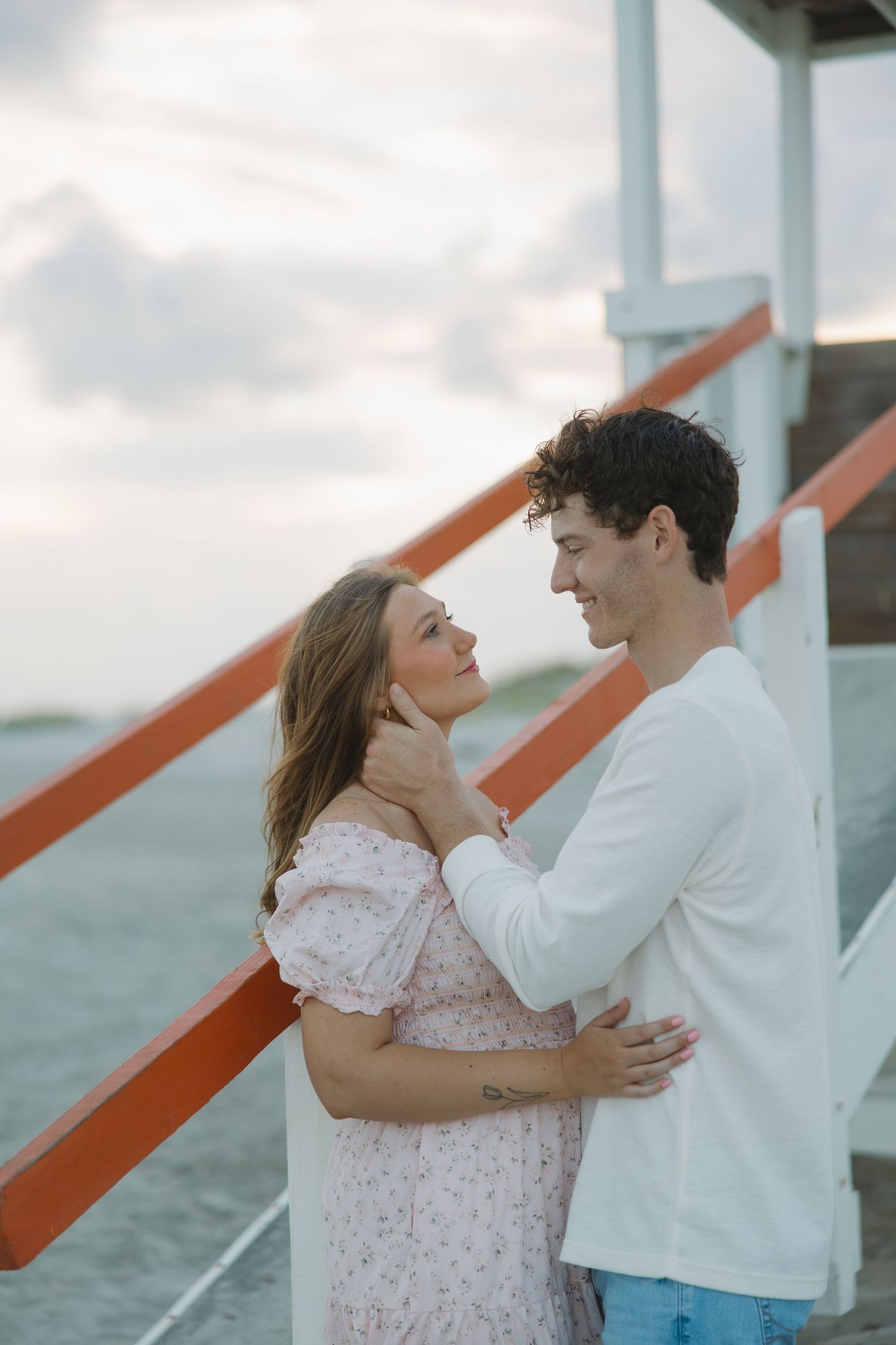 A couple embraces romantically near a beach lifeguard tower, smiling at each other at sunset.