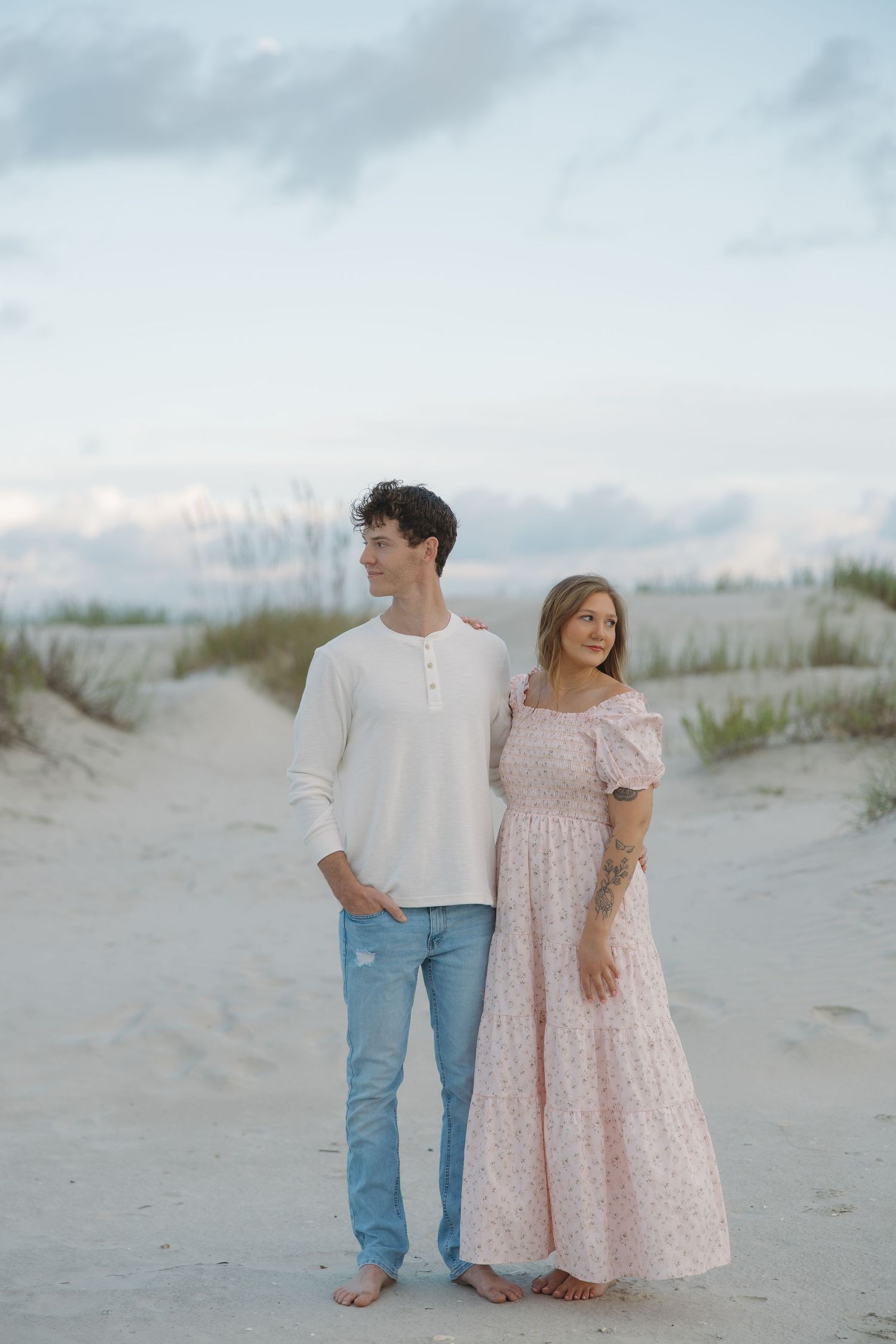 A couple stands together on a sandy beach path, man in white shirt and jeans, woman in floral pink maxi dress.