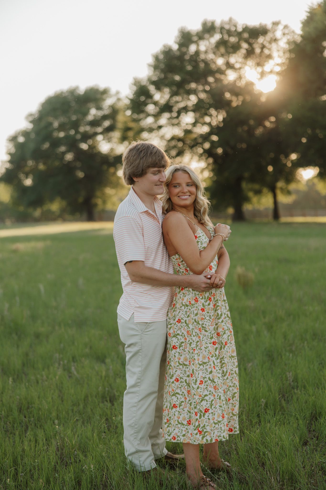 A couple embraces in a sunlit green field at golden hour, the woman wearing a floral dress smiling warmly.