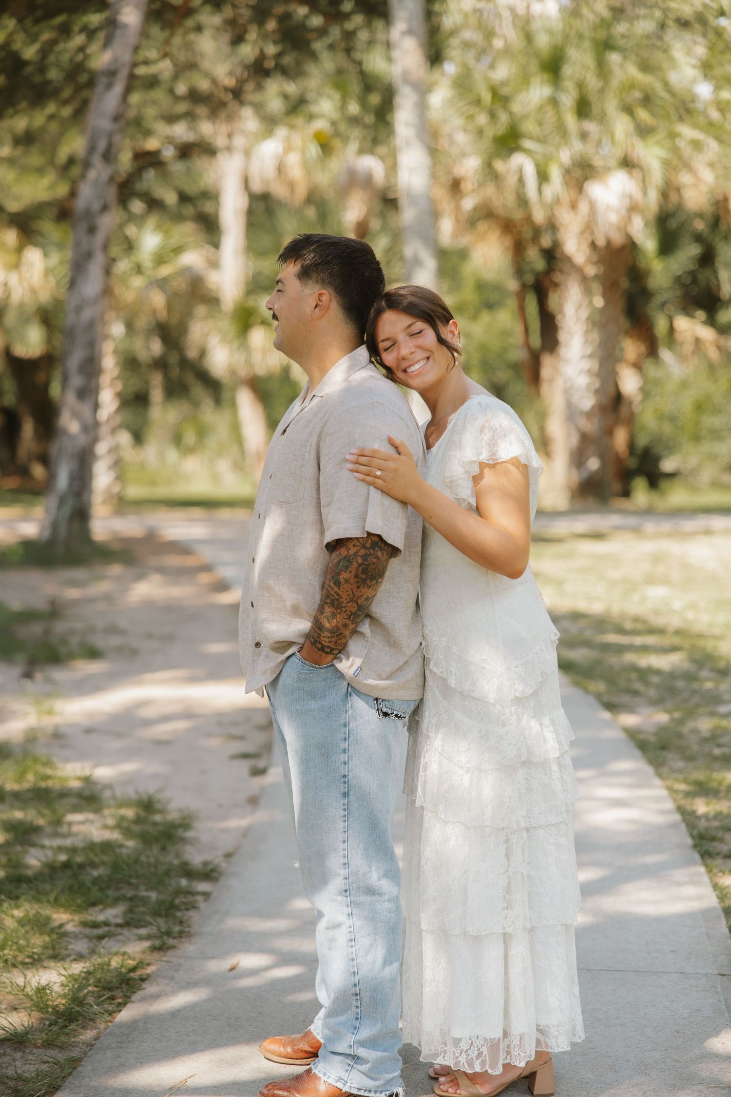 Couple embracing on a sunny path, woman in white tiered dress leaning on mans shoulder, surrounded by palm trees.