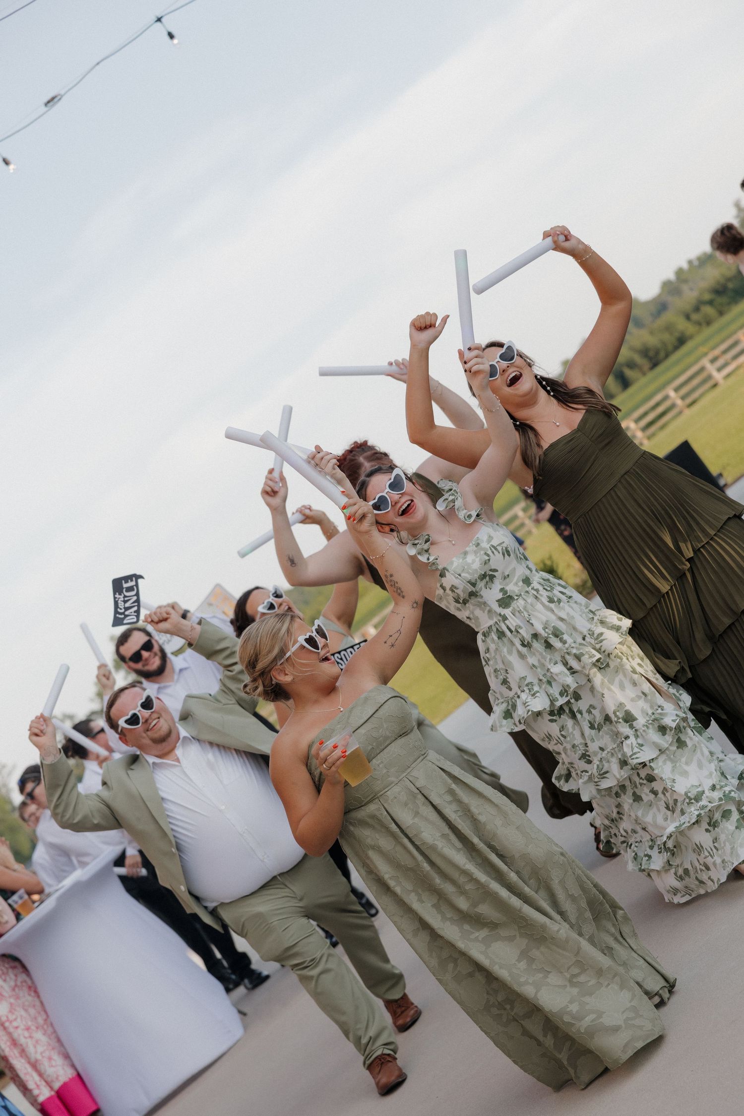 Wedding guests in sage green dresses and sunglasses cheer and dance outdoors while waving white glow sticks at an evening reception.