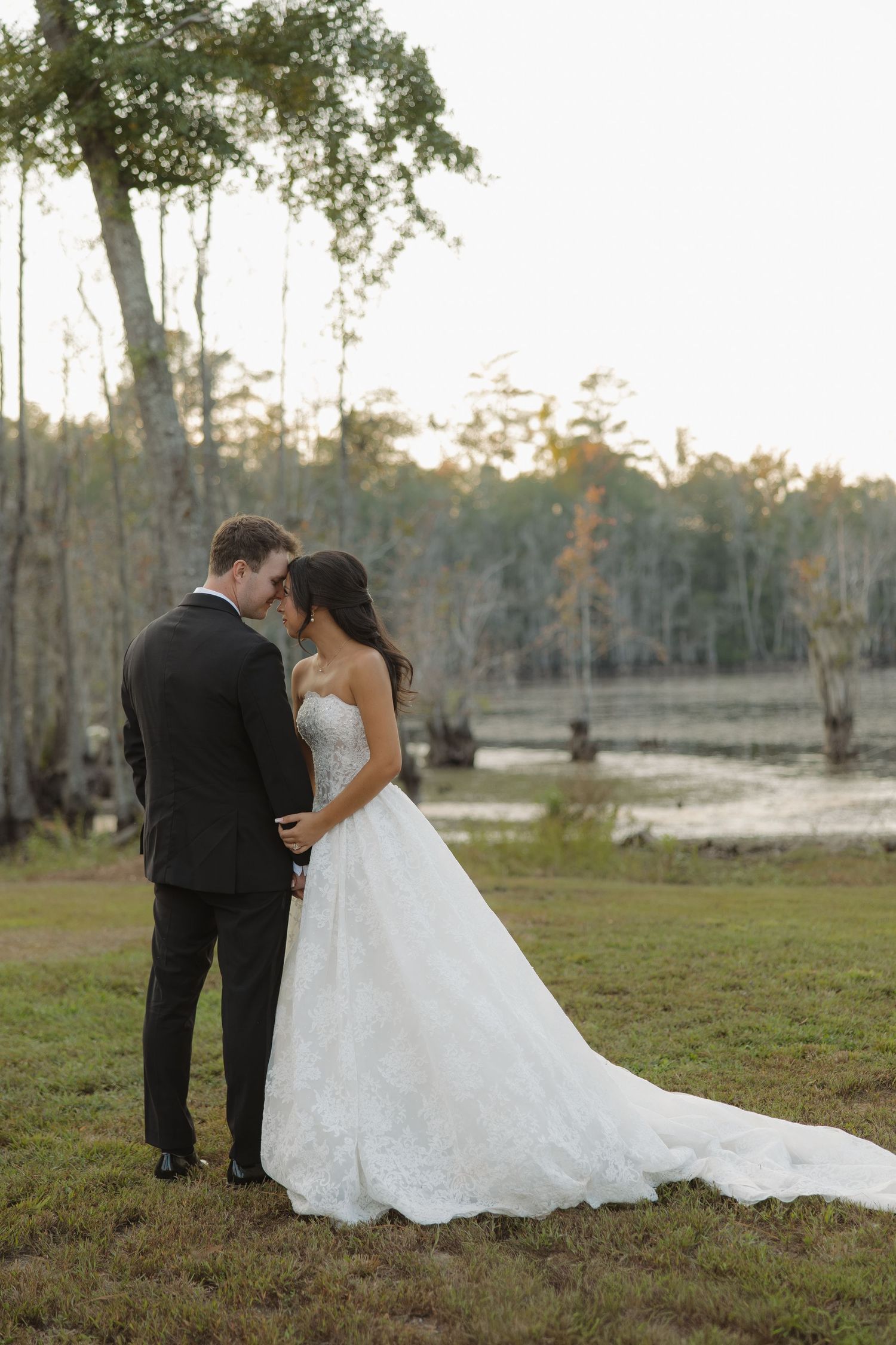 Bride in a strapless lace wedding gown and groom in a black suit touch foreheads on a grassy lawn beside a misty cypress swamp.