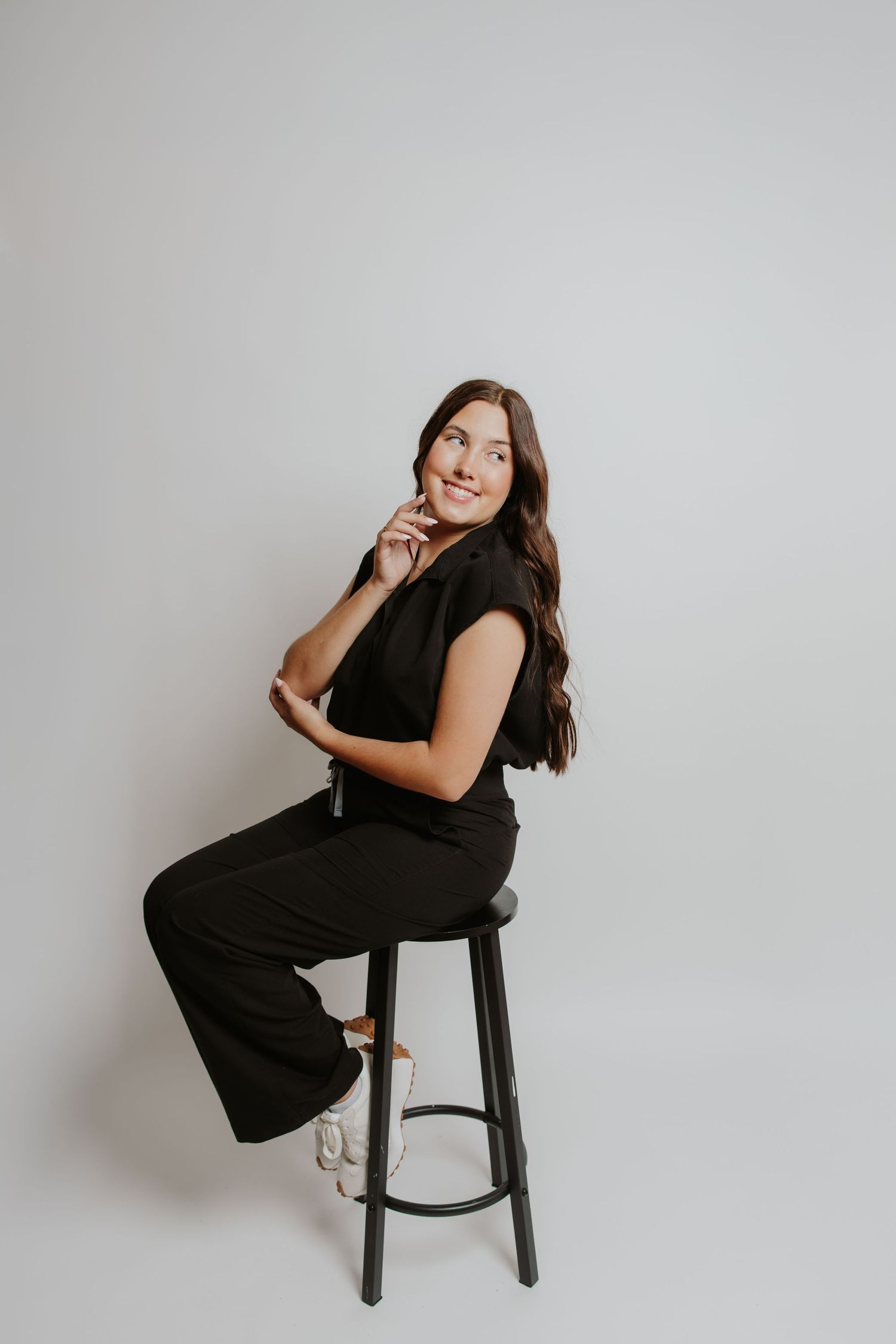 Young woman in black outfit sitting on a stool, smiling thoughtfully against a white background.