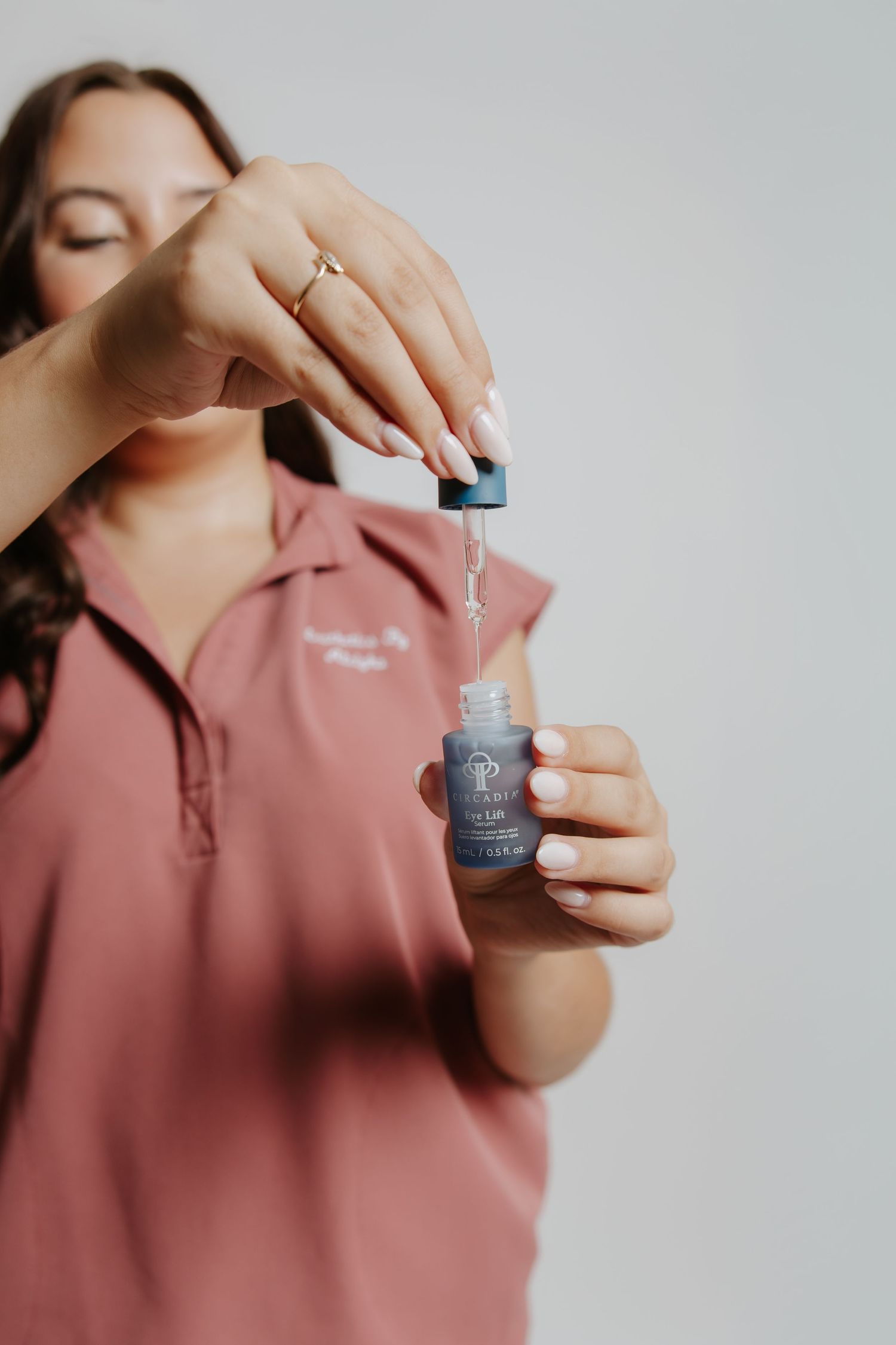 Woman in pink top holding a dropper bottle of serum, applying drops with manicured hands against white background.