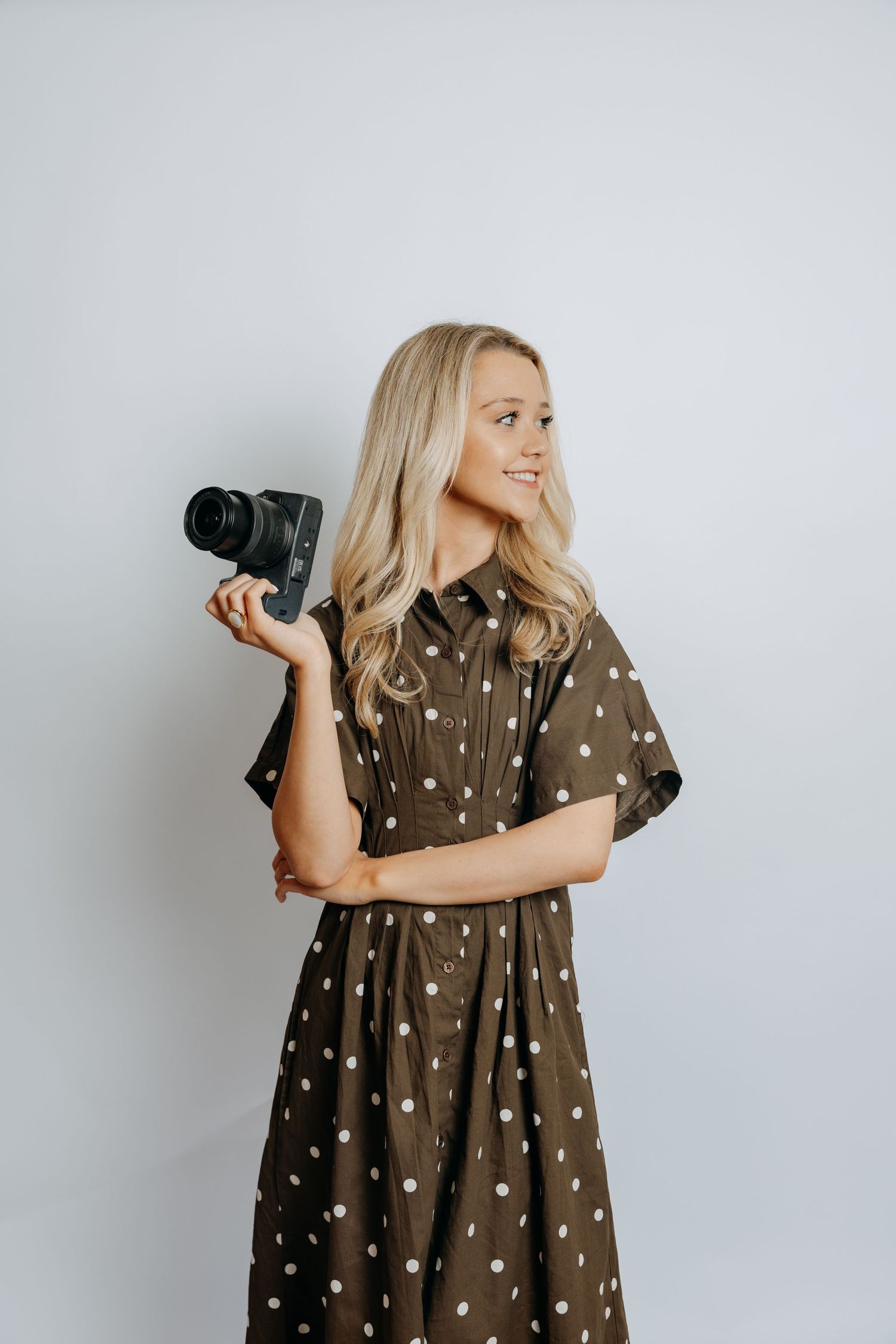 Blonde woman in brown polka dot dress smiling while holding a DSLR camera.