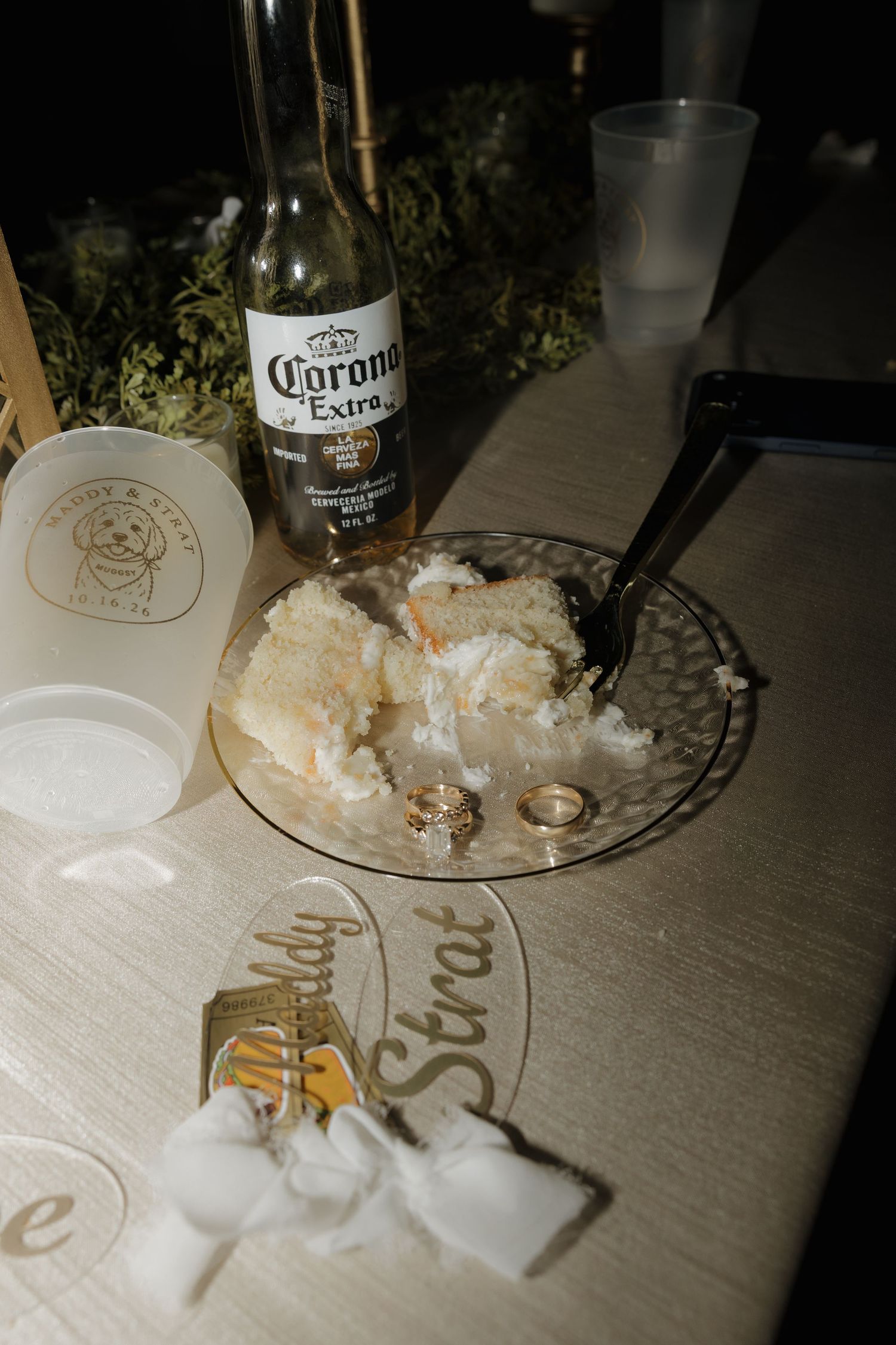 Two wedding rings rest on a plate of leftover cake beside a Corona Extra bottle, cups, and personalized name place cards on a table.