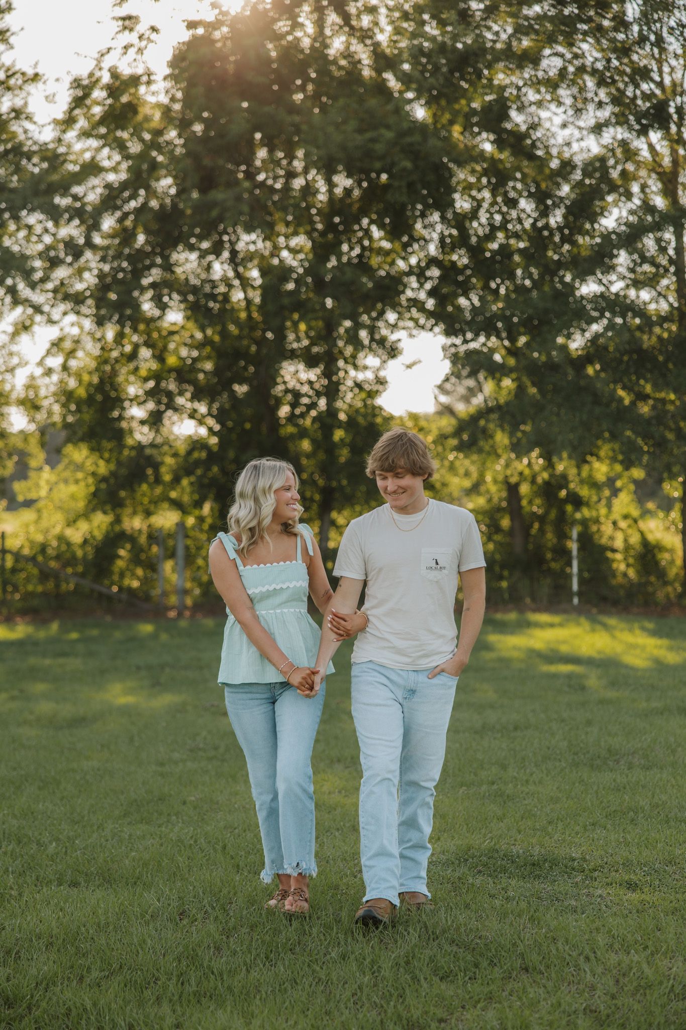 A young couple holds hands while walking on green grass at golden hour with trees in the background.