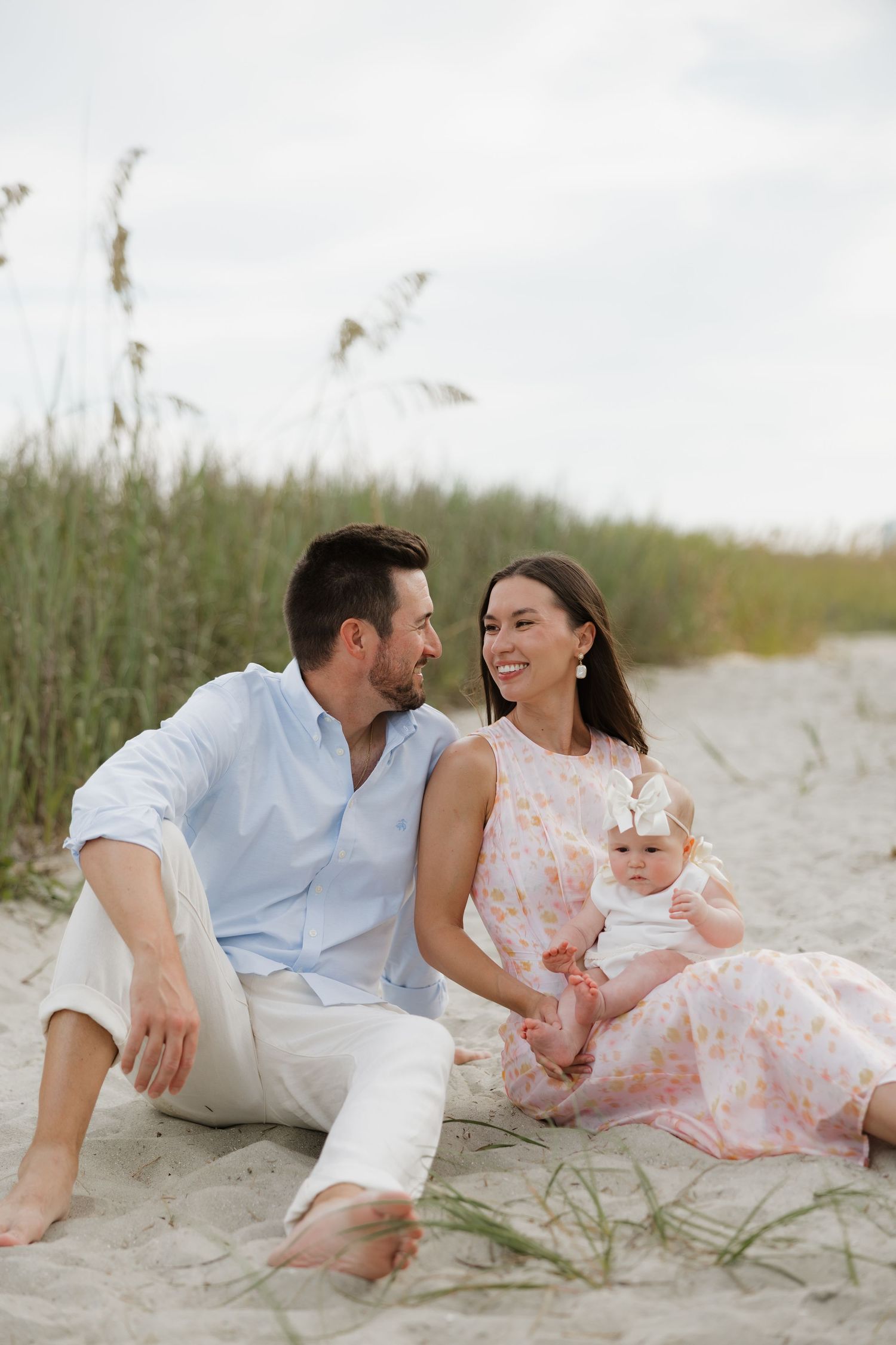 A happy young family with a baby girl sits on sandy beach dunes with sea oats in the background.