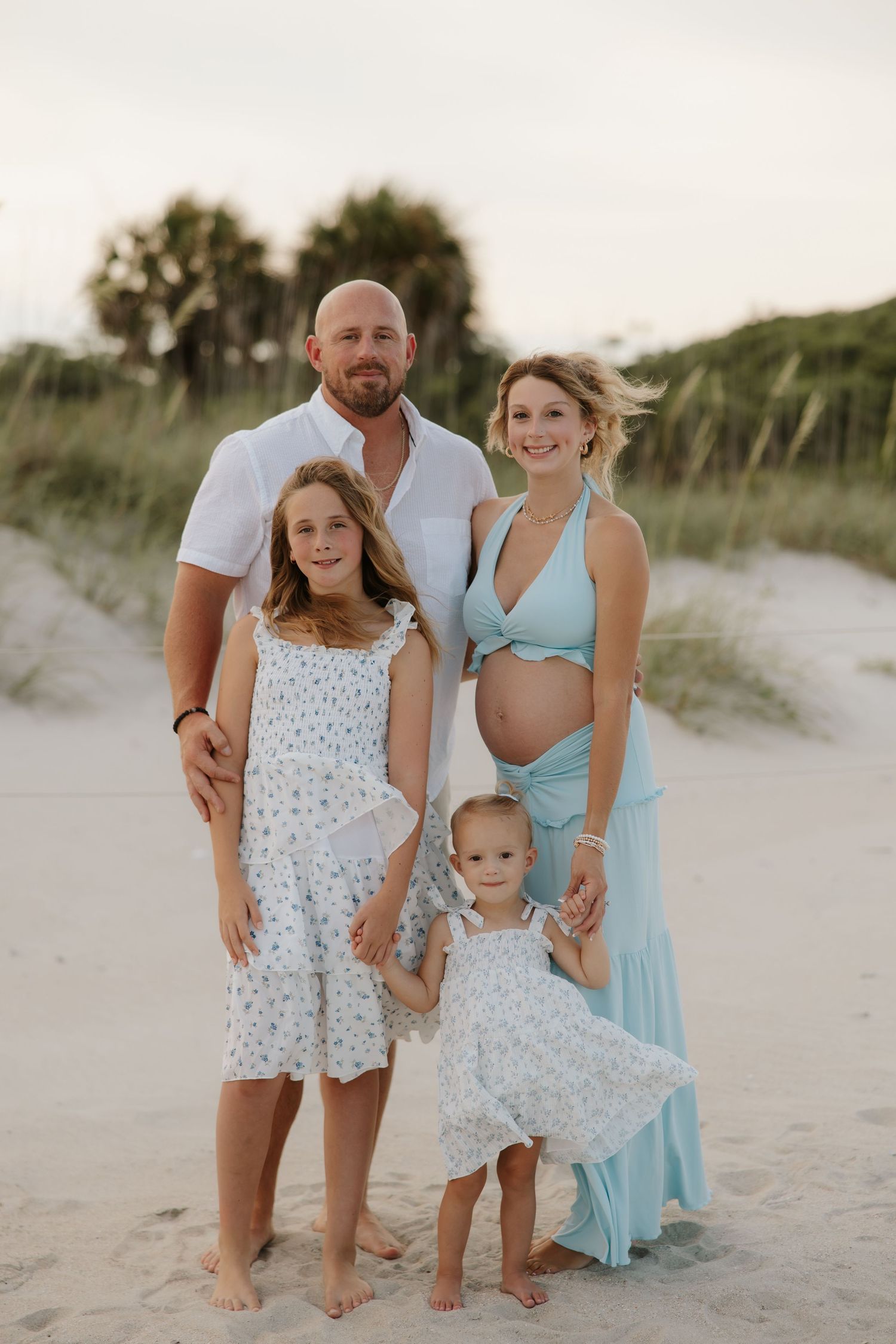 Pregnant woman in teal dress poses with husband and two daughters for a family beach maternity photo session.