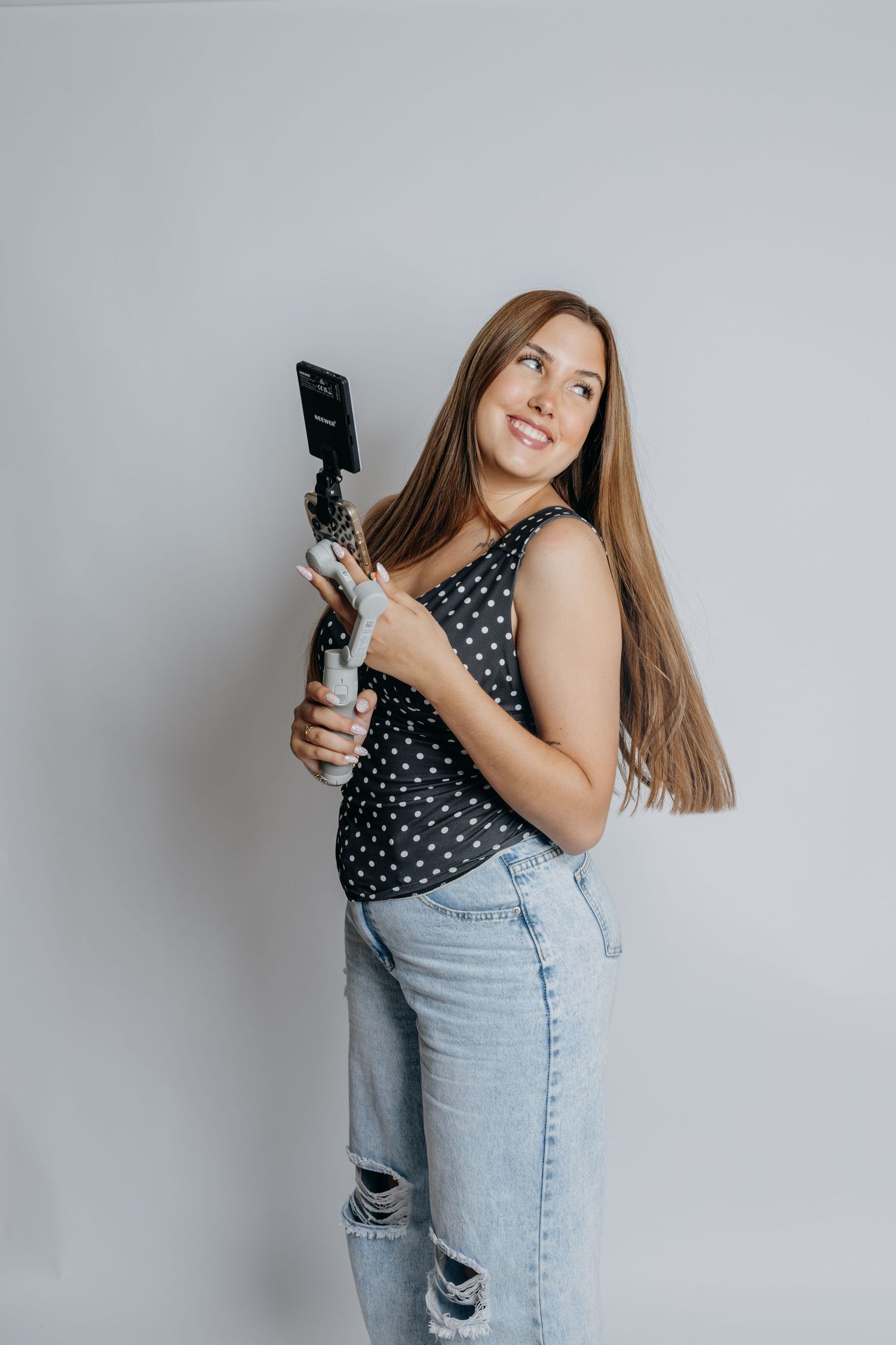 Smiling woman in polka dot tank top holding smartphone gimbal stabilizer against light gray background.