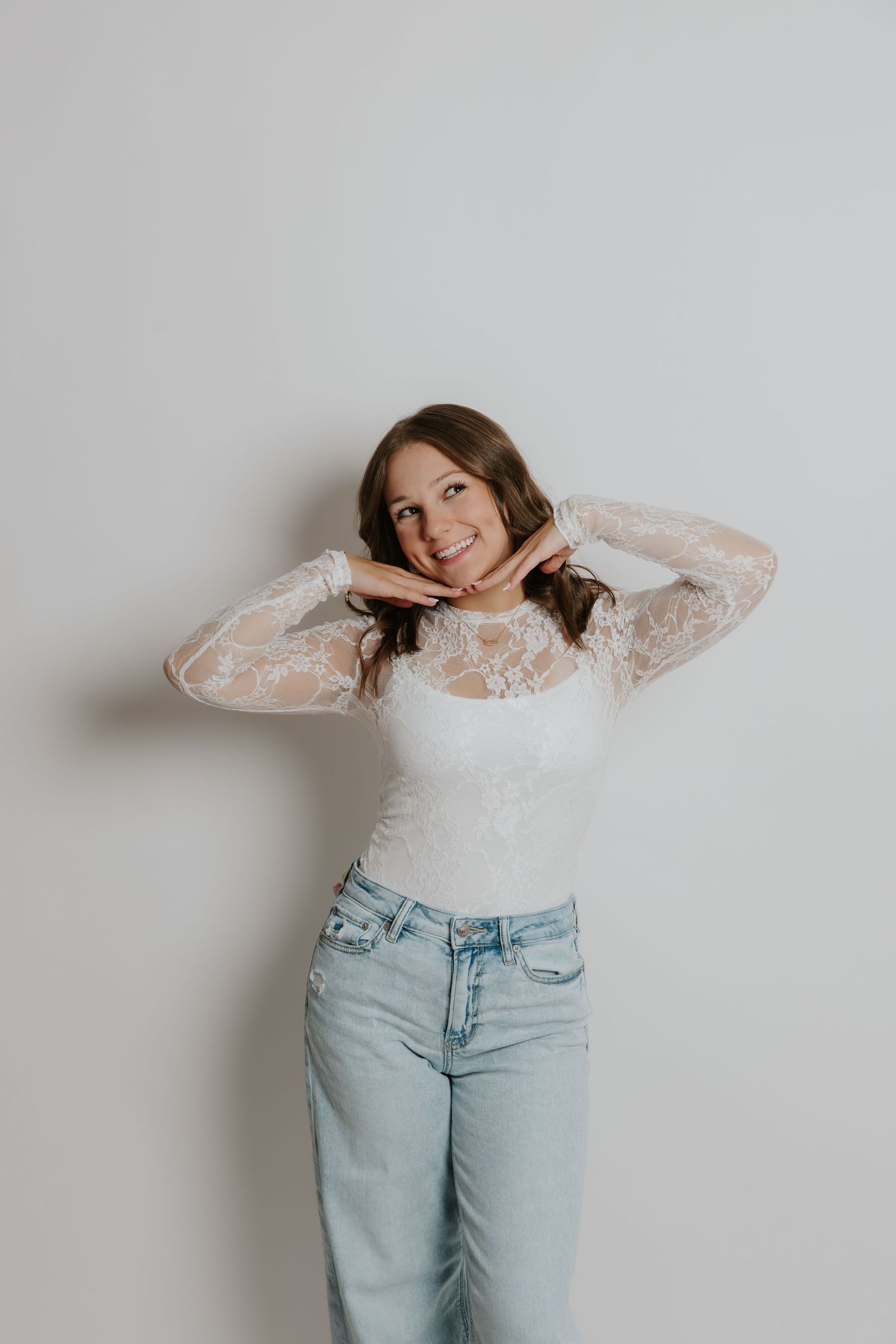 Smiling young woman in white lace long sleeve top and light wash jeans posing against white wall.