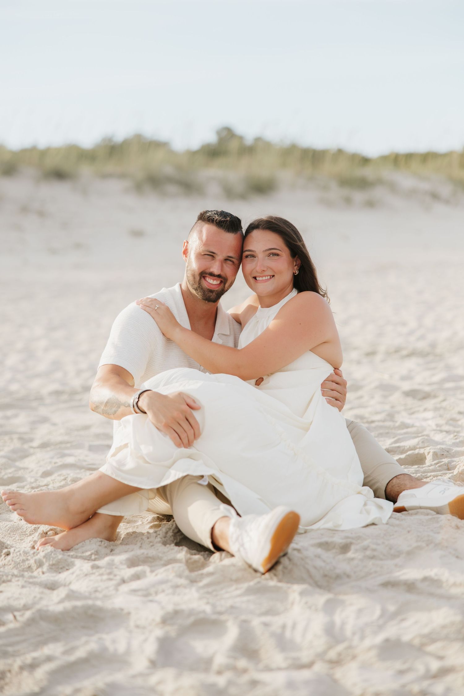 Happy couple sitting together on sandy beach, man embracing woman in white dress, smiling at camera.