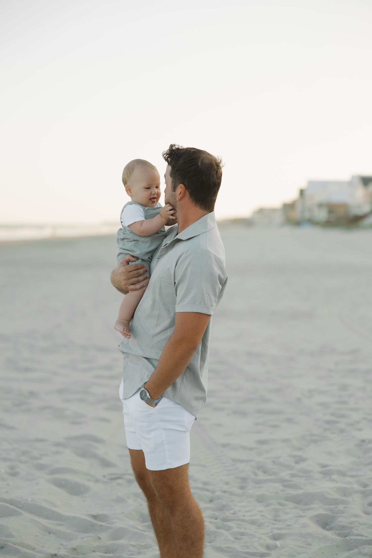A father holds his baby on a sandy beach at sunset, both dressed in light blue and white outfits.