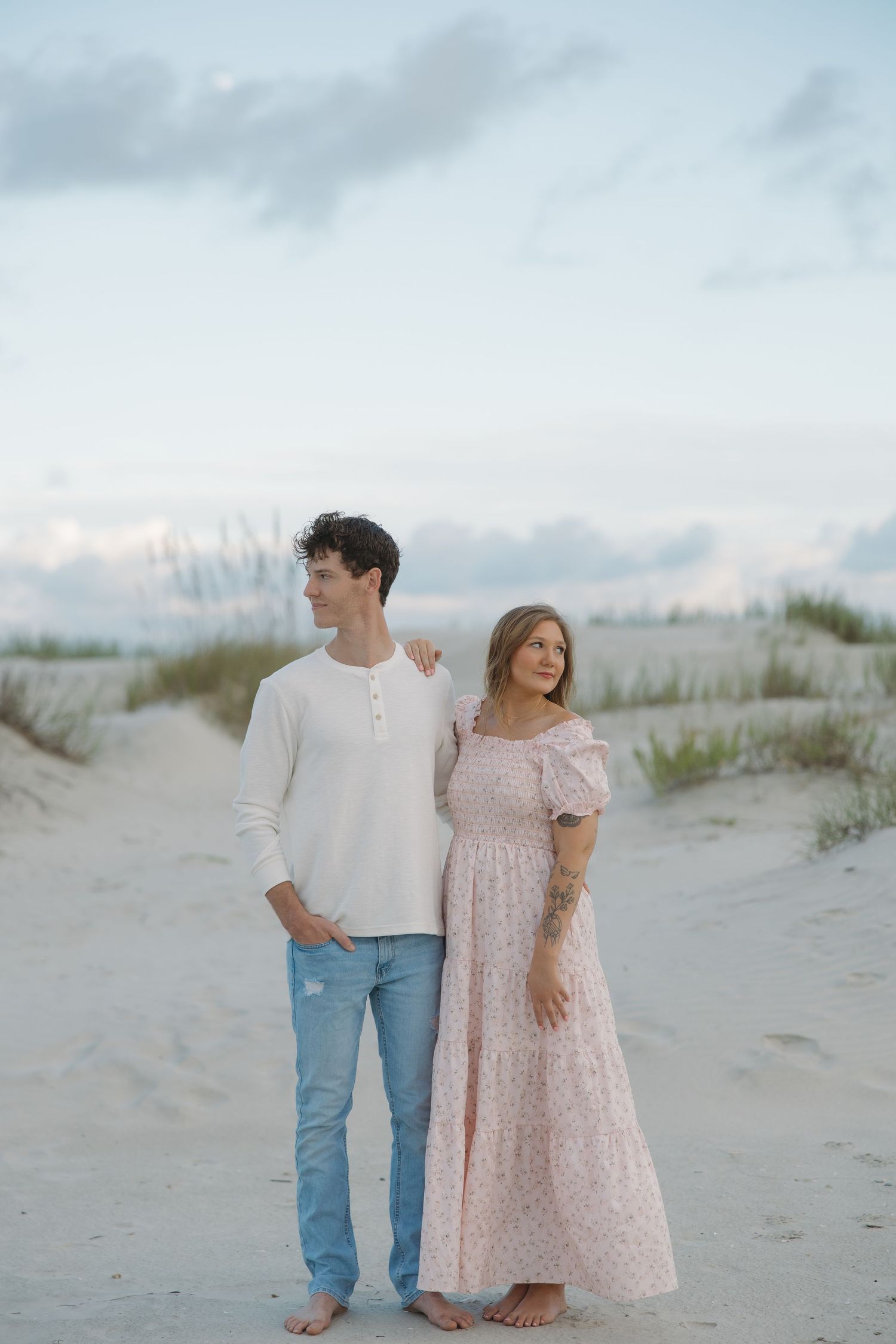 A young man and woman stand together on a sandy beach, wearing casual pastel outfits, with dunes and cloudy sky behind them.
