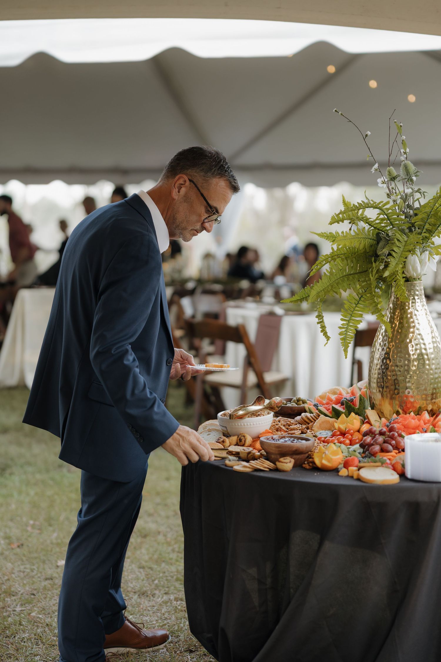 A man in a navy suit and glasses serves himself from a wedding buffet of fruit, bread and snacks under a white tent.