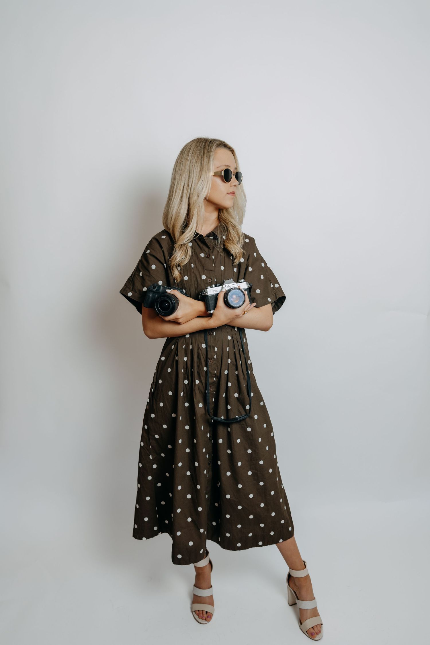 Blonde woman in an olive polka dot midi dress holding cameras while wearing sunglasses and heels.