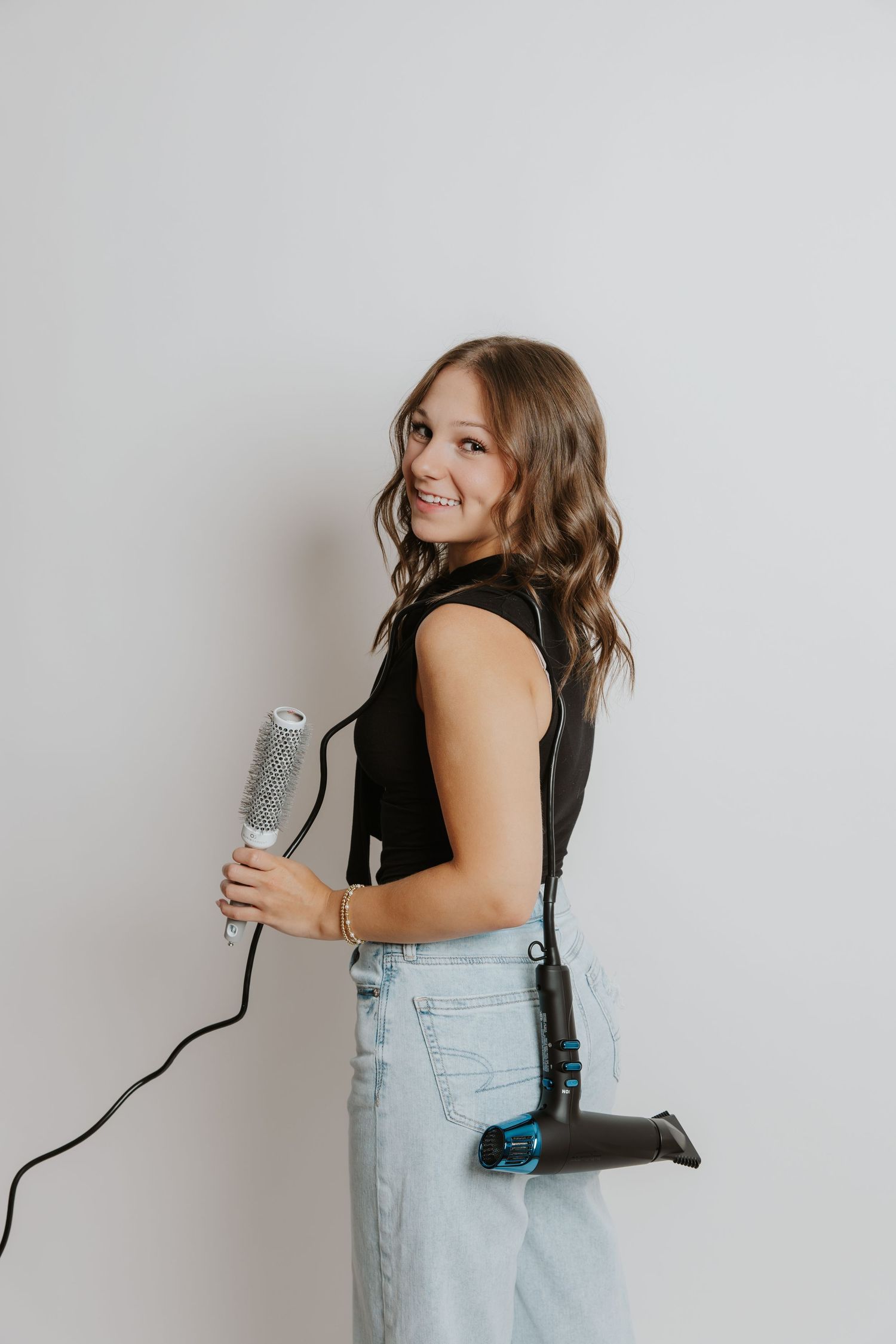 Smiling woman in black top holds round brush with hairdryer tucked in jeans against white wall.