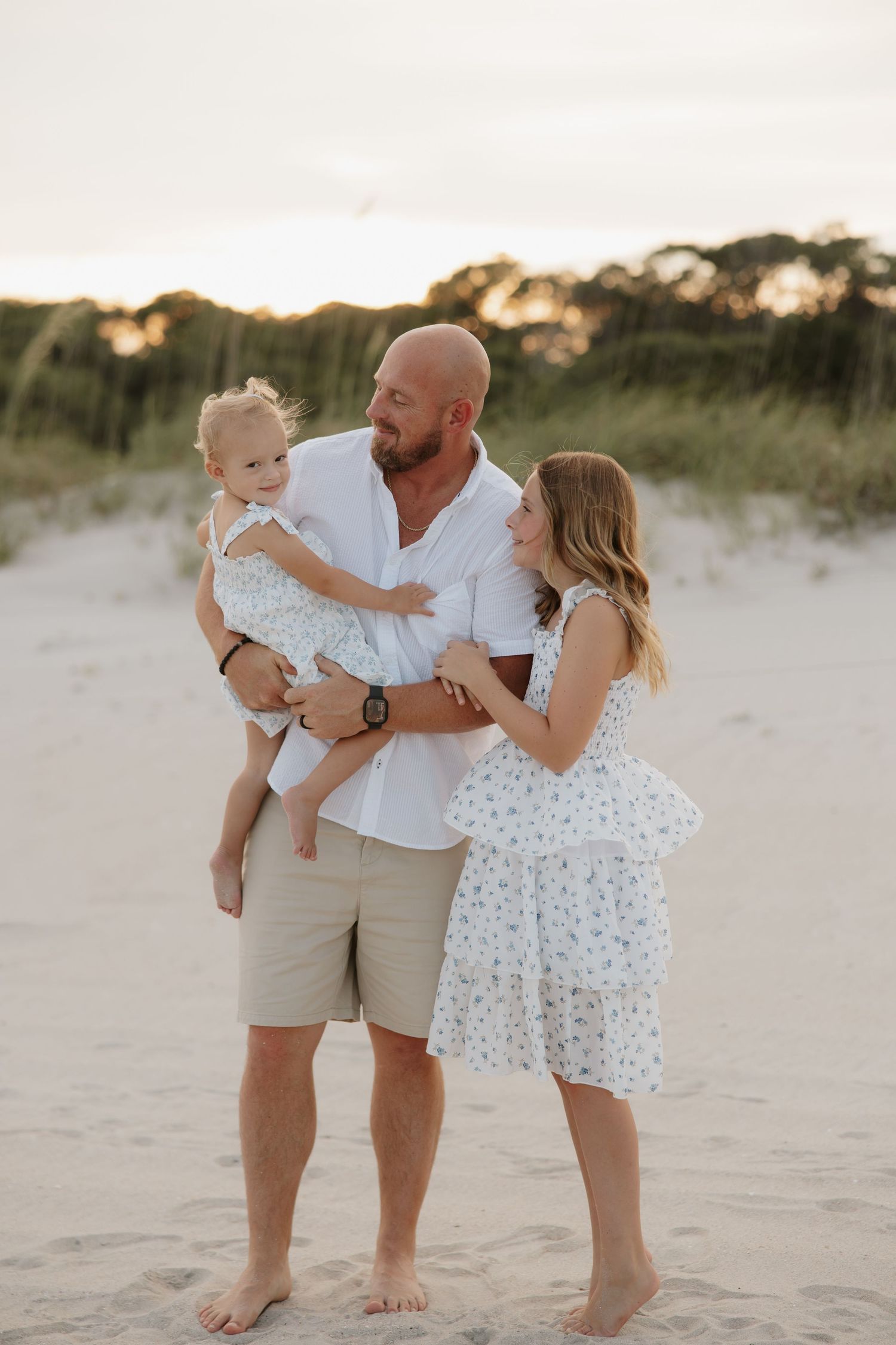 A family of three poses on a sandy beach at sunset, with a bald man holding a toddler while a woman in a floral dress looks on.