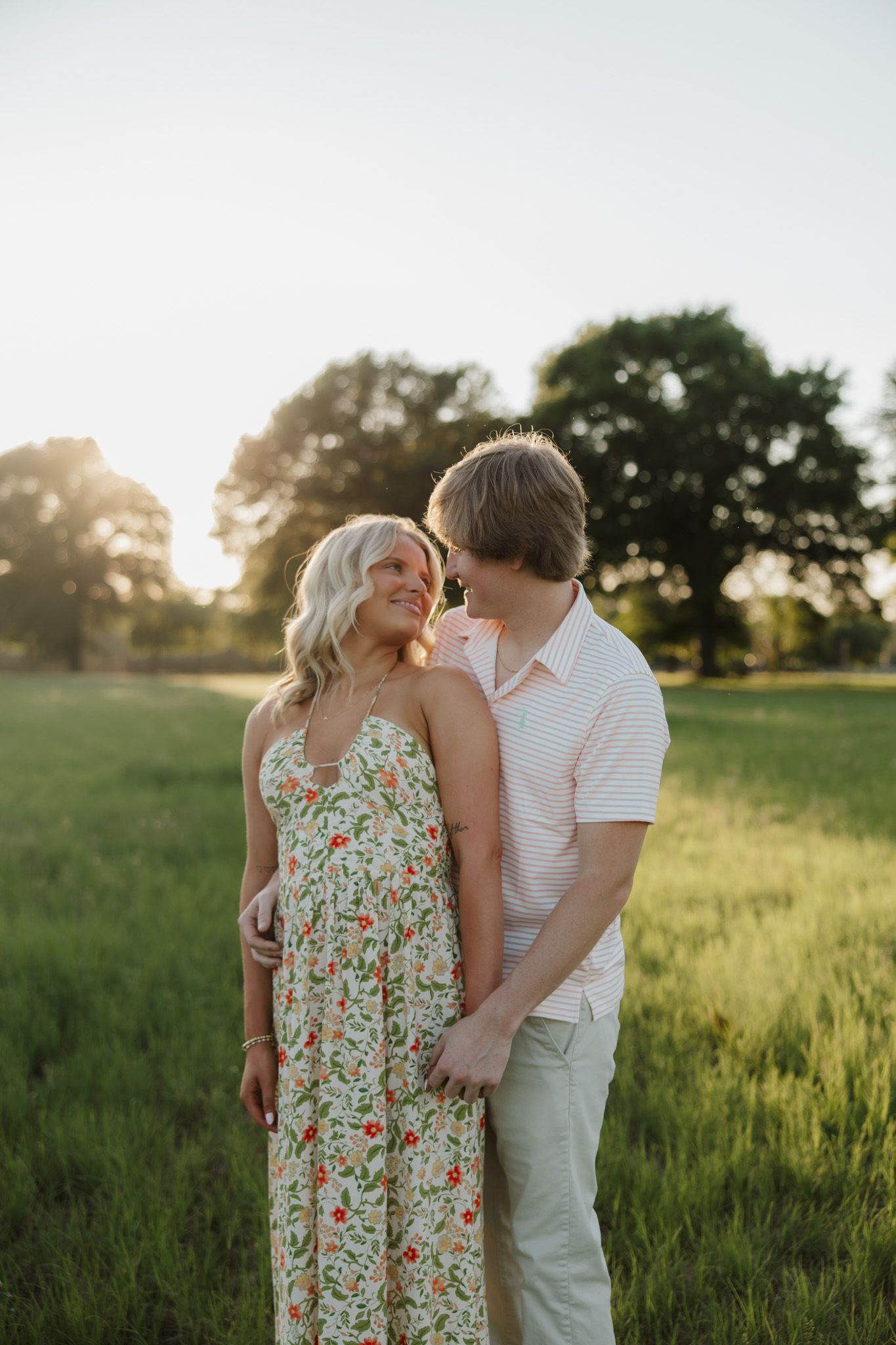 Couple embracing in a sunlit green field at golden hour, woman in floral maxi dress smiling up at man in pink polo.