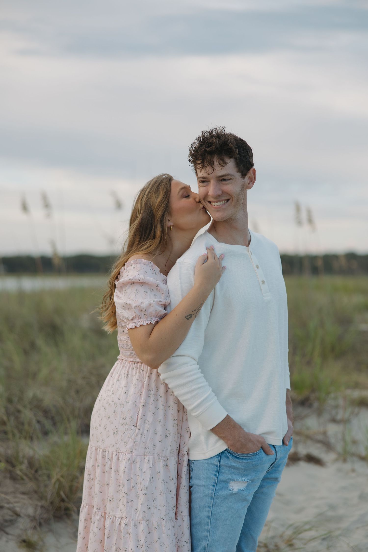 Woman in pink floral dress kissing smiling mans cheek on a sandy beach with sea oats behind them.