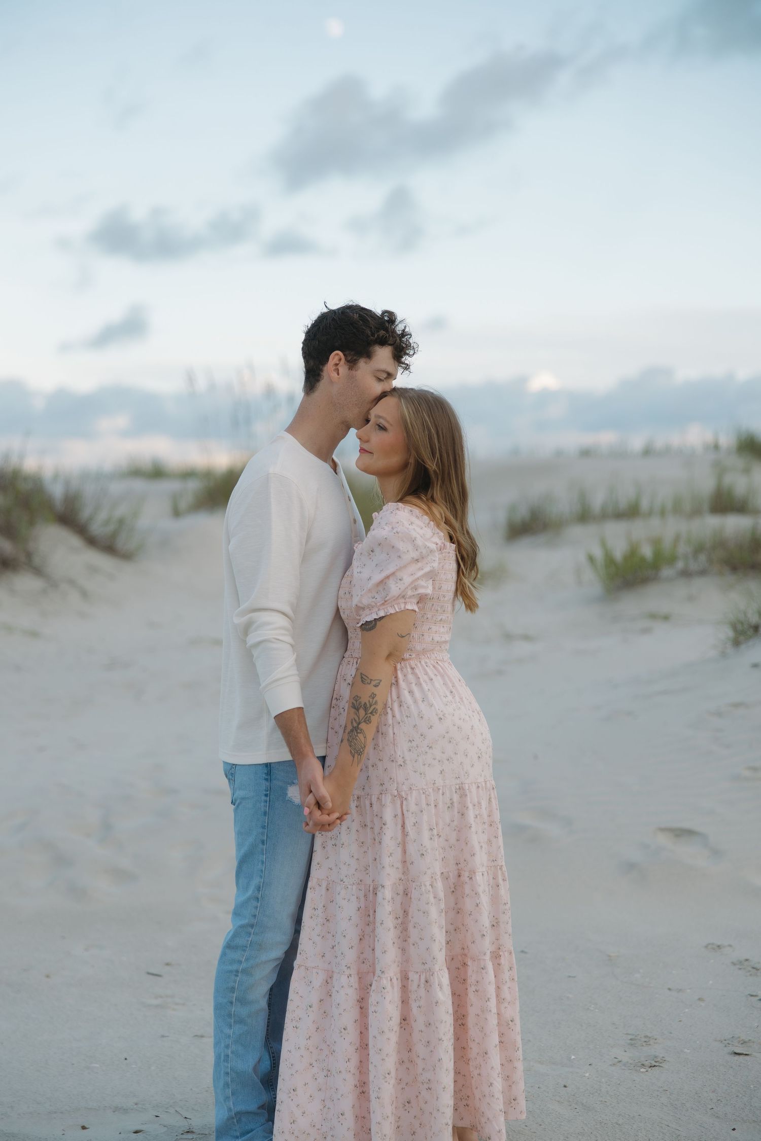 A couple holds hands and shares a tender moment on a sandy beach at dusk, woman in a floral pink maxi dress.
